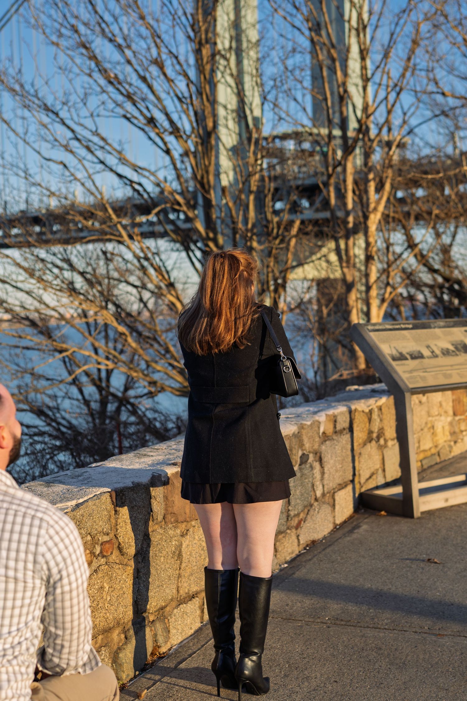 Young woman in black outfit standing near a stone wall with a suspension bridge backdrop.
