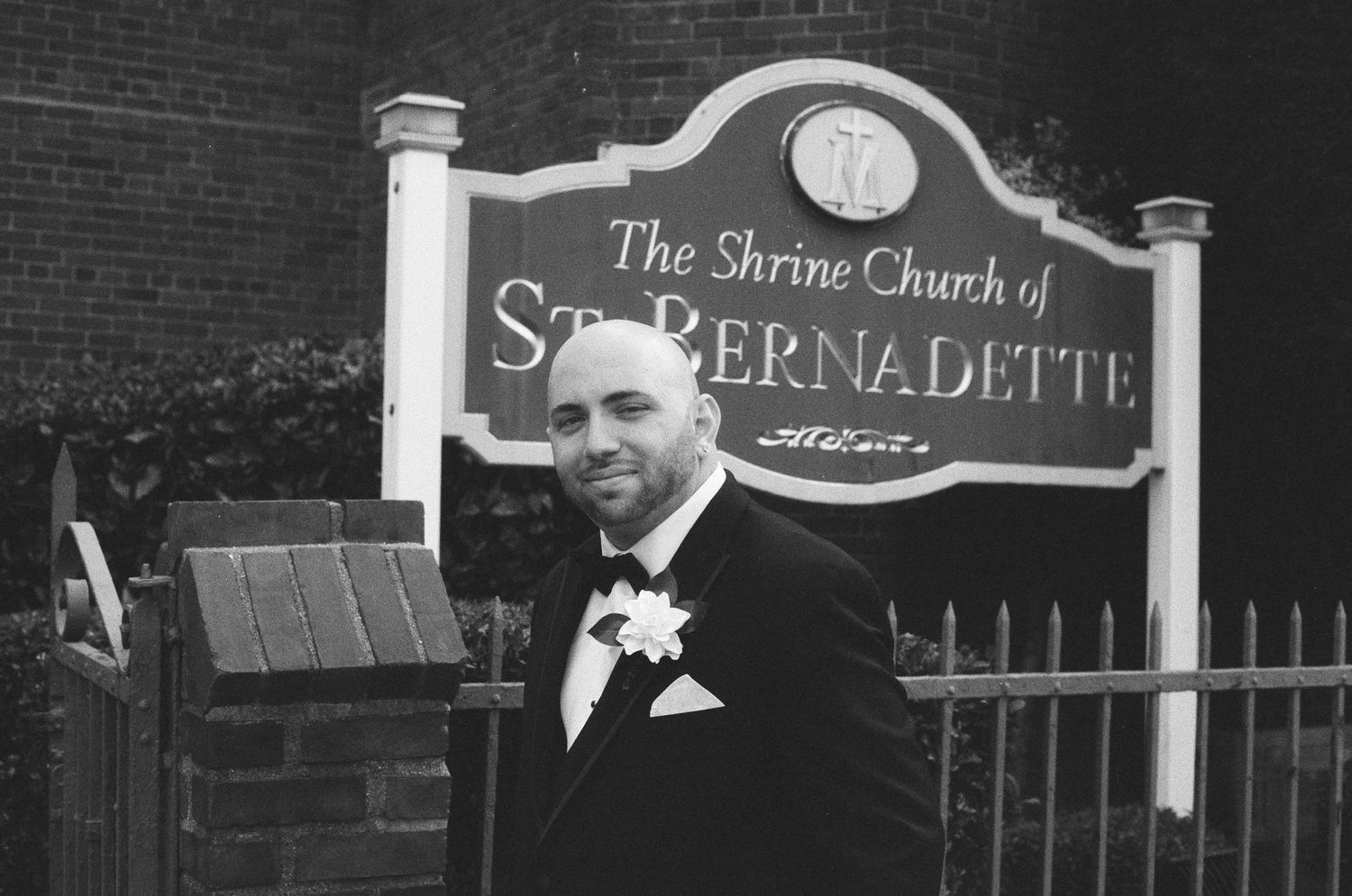 Groom in tuxedo smiling beside The Shrine Church of St. Bernadette sign in black and white.