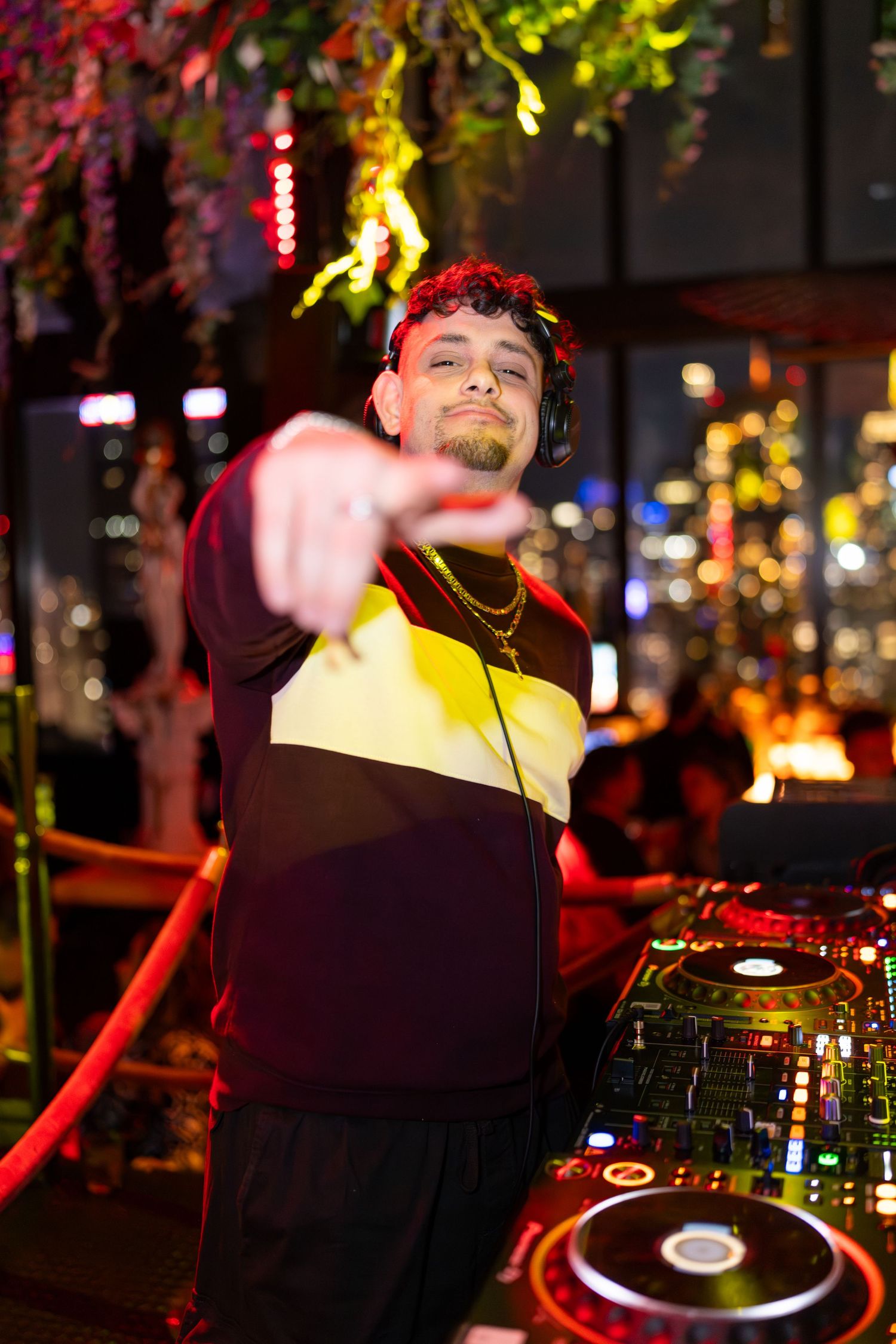 DJ pointing at camera while mixing at colorful nightclub event with city lights in background.