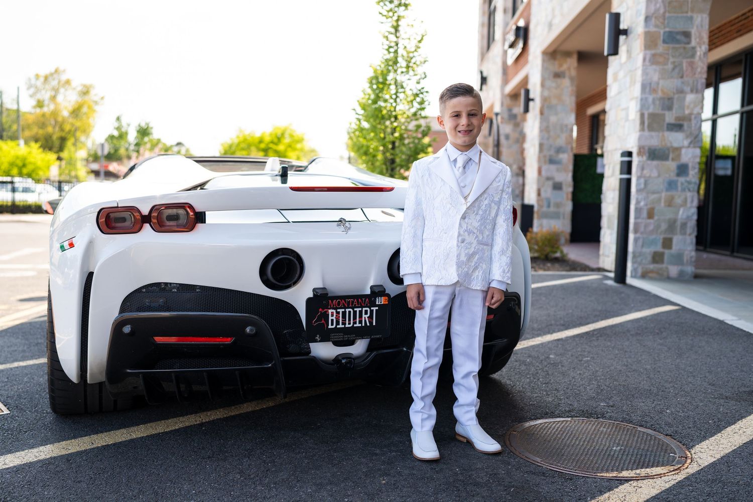 Young boy in white suit posing beside a white sports car with BIDIRT license plate in a parking lot.