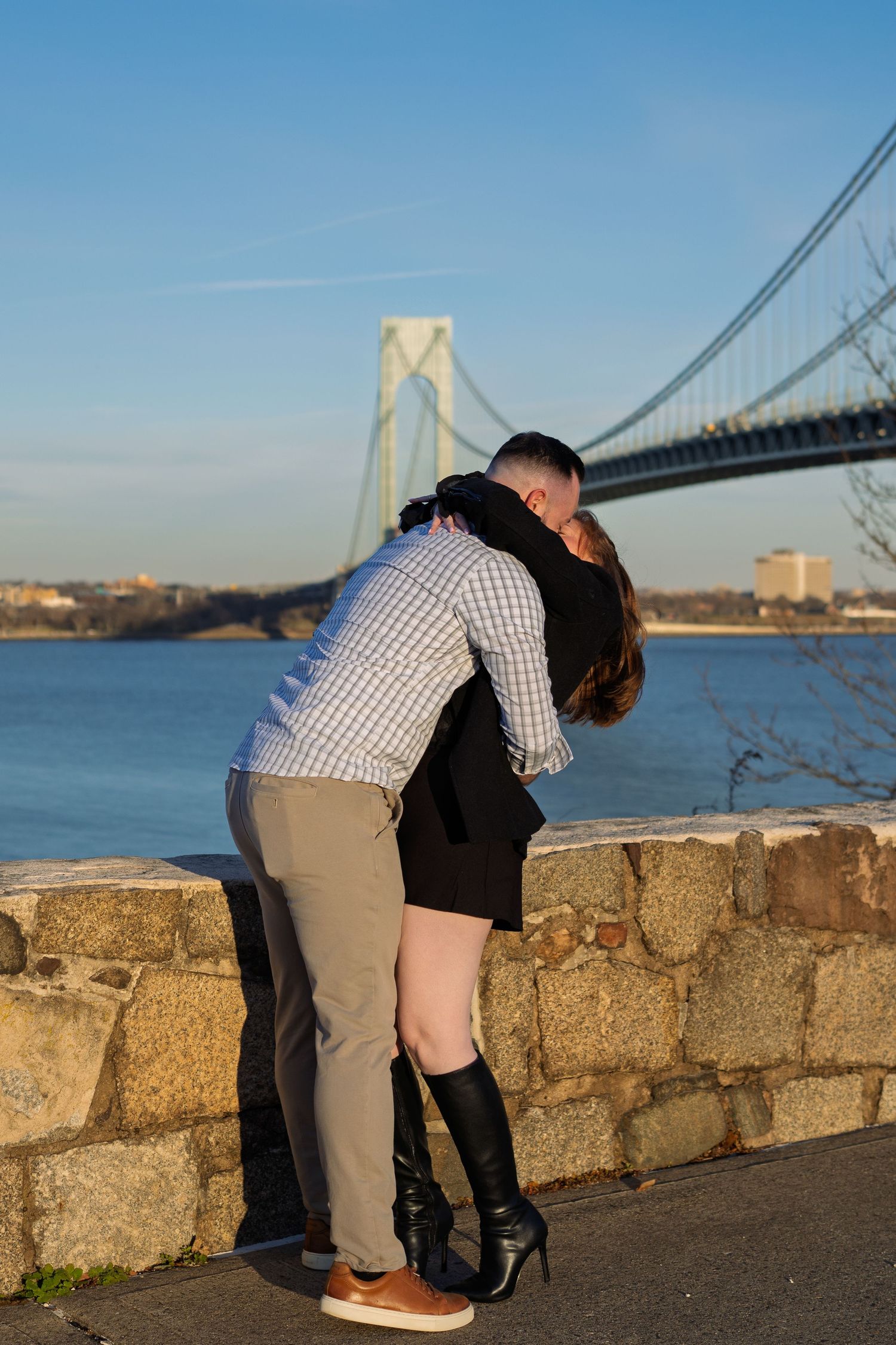 Couple sharing a romantic embrace by a stone wall with the Verrazzano-Narrows Bridge in the background.