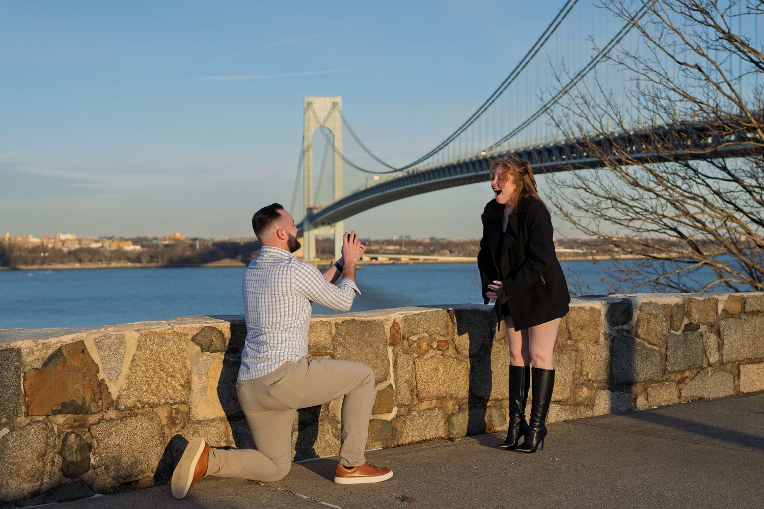 Couple at romantic marriage proposal by waterfront with suspension bridge backdrop on sunny day.