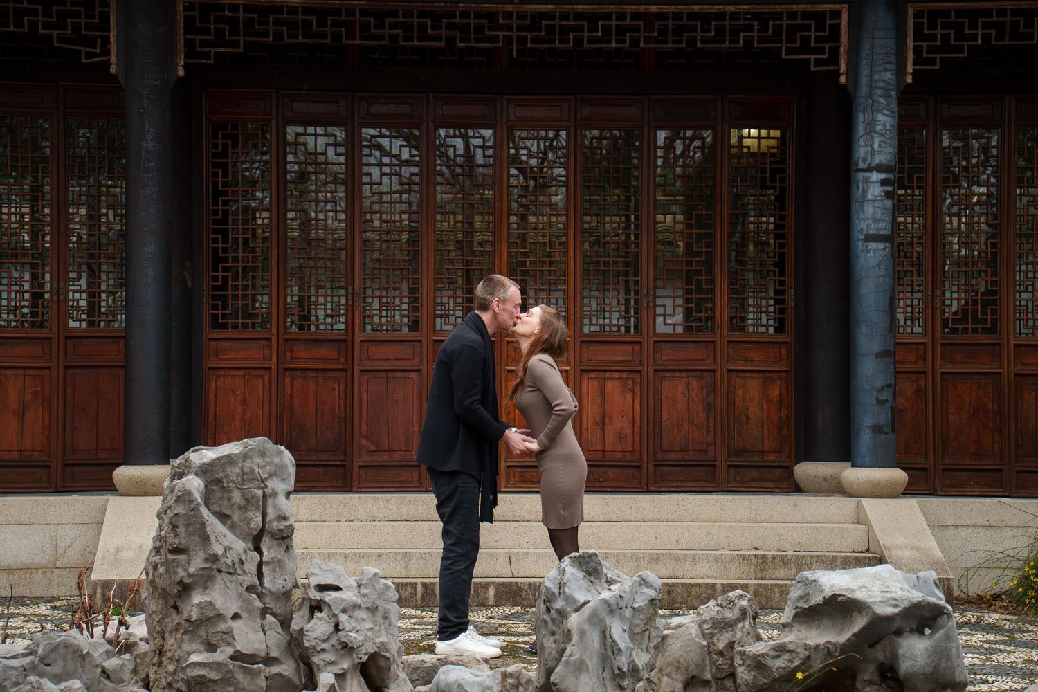 Couple sharing a tender kiss in front of ornate wooden doors of a Chinese pavilion.