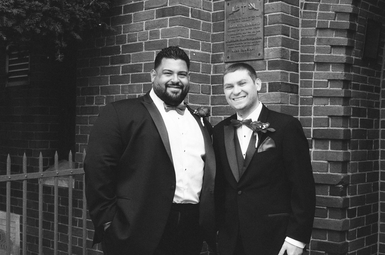 Black and white photo of two groomsmen in tuxedos smiling in front of a brick wall.