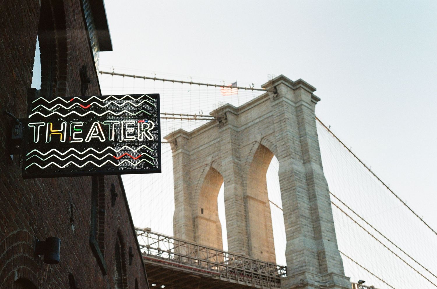A neon Theater sign mounted on a brick wall with the Brooklyn Bridge tower in the background.