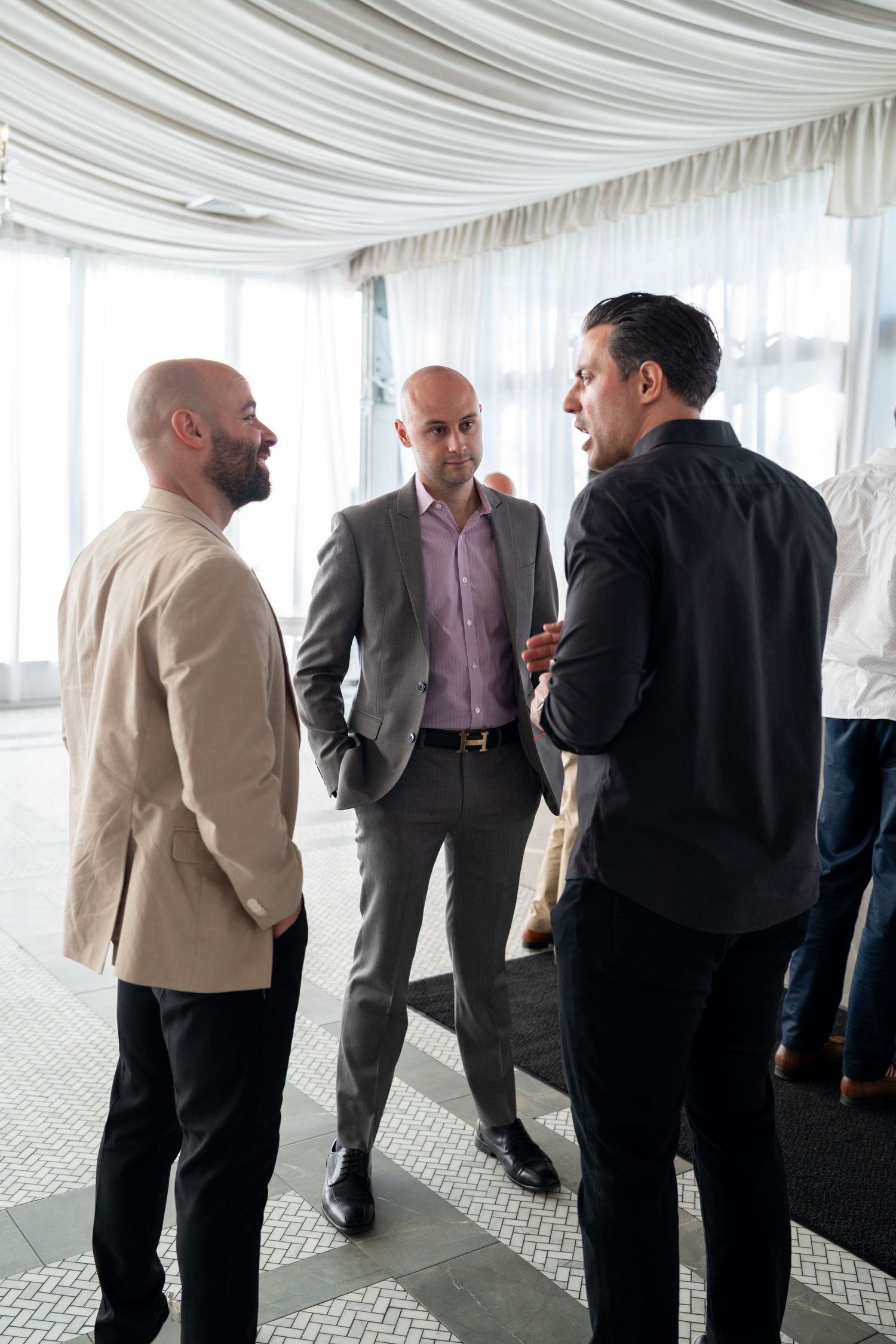 Three bald men in business attire engaged in conversation at an elegant white draped venue.