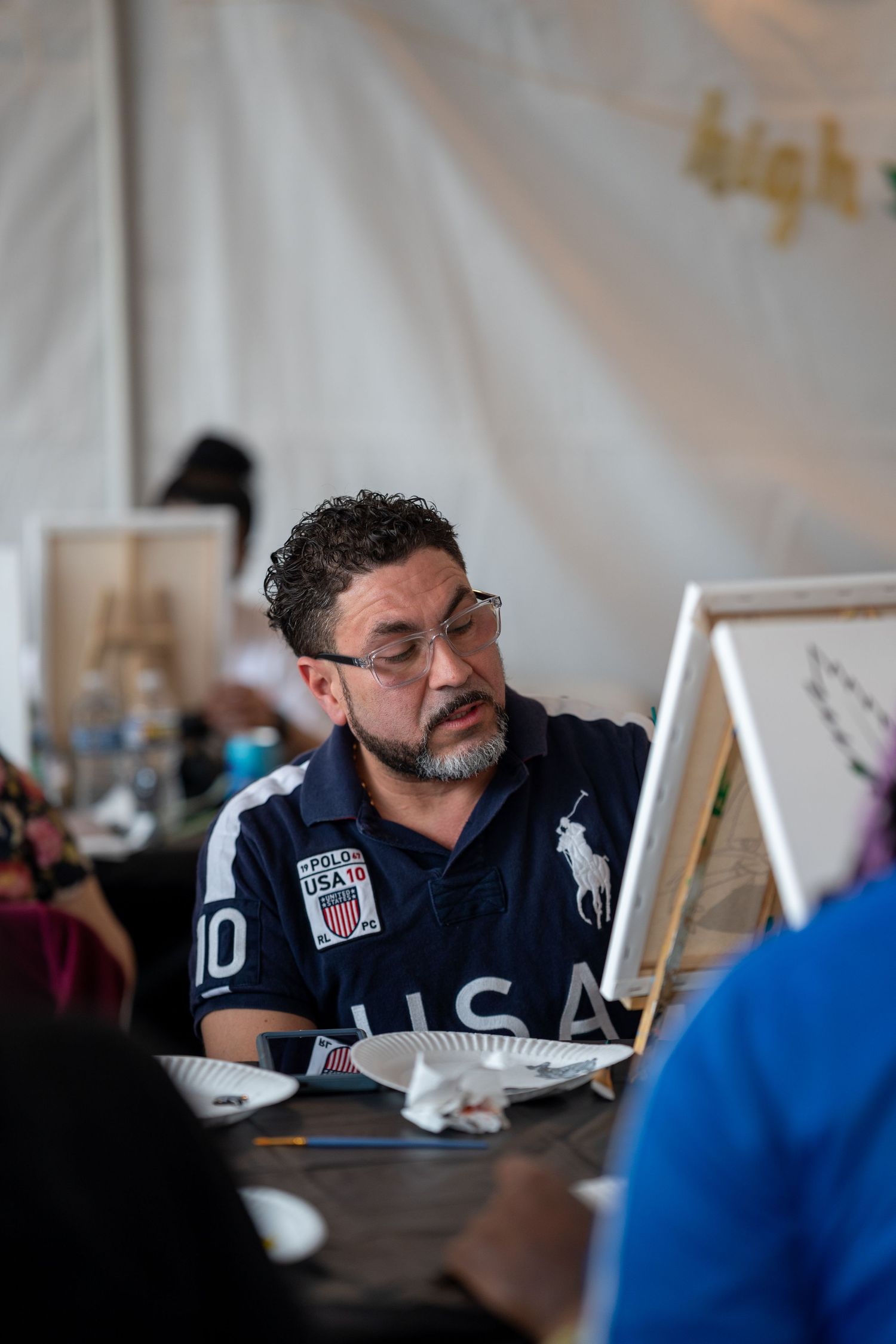 Man in USA polo shirt focused on painting at canvas during outdoor art event.