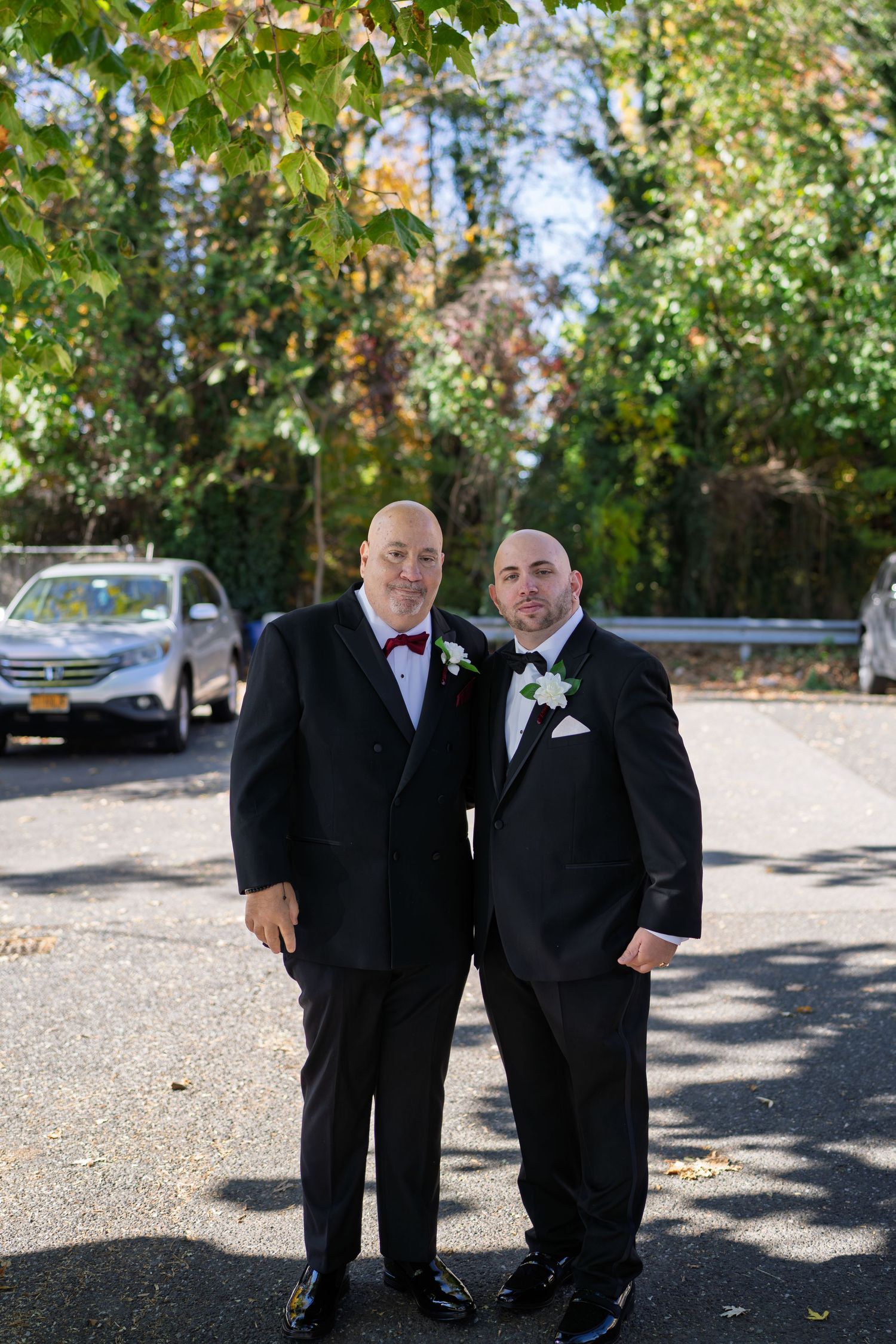 Two men in black tuxedos with boutonnieres pose together outdoors at a fall wedding.