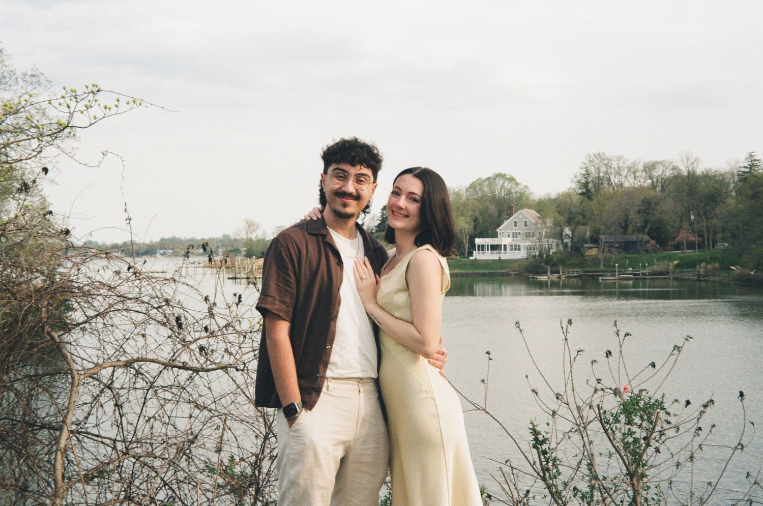 Smiling couple standing close together near a lake with bare branches and houses in the background.