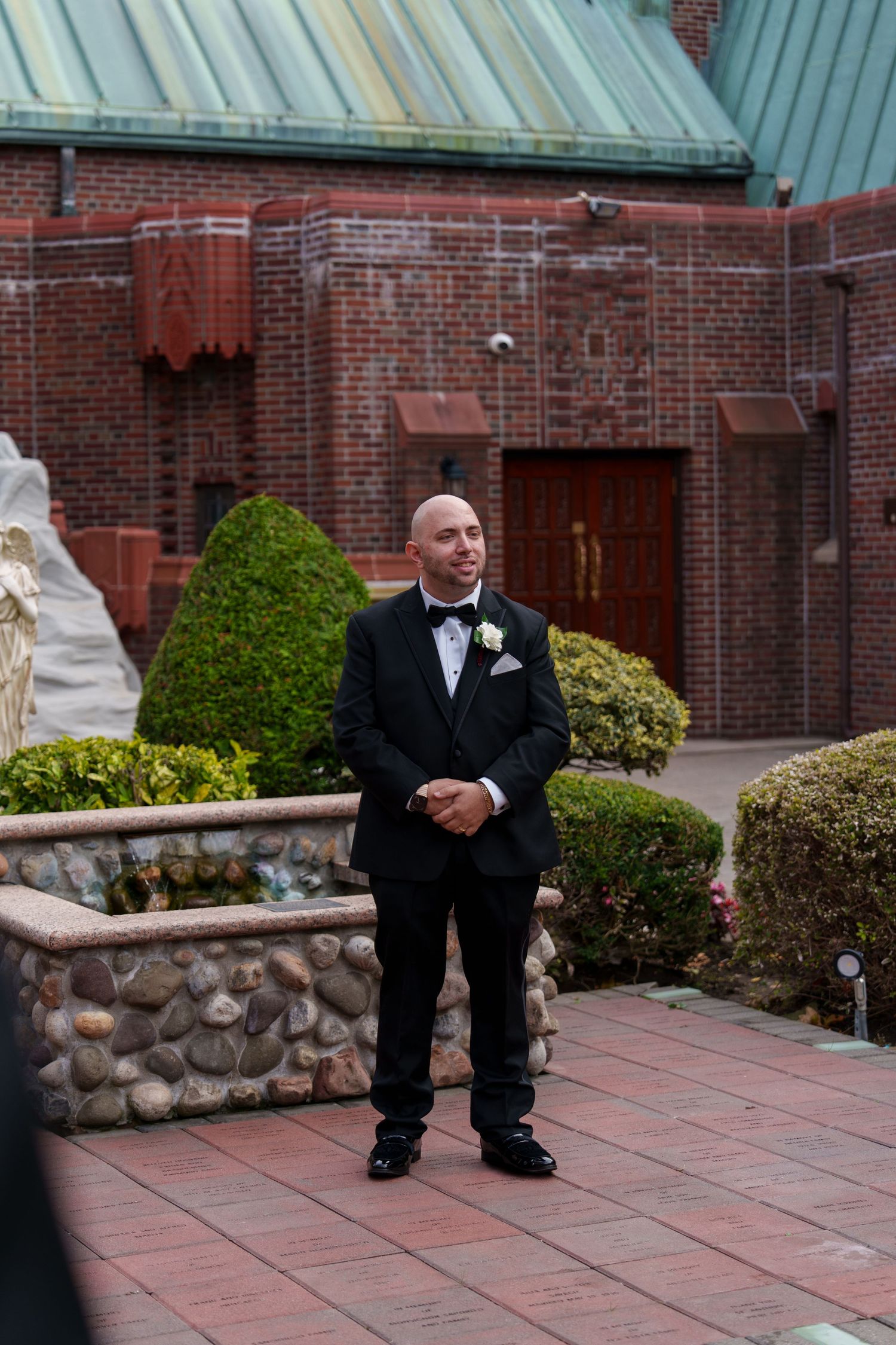 Groom in tuxedo waiting outside brick church with manicured gardens before wedding ceremony.