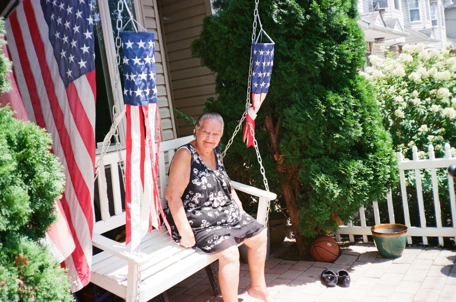 Elderly person sitting on porch swing surrounded by American flags on a sunny day.