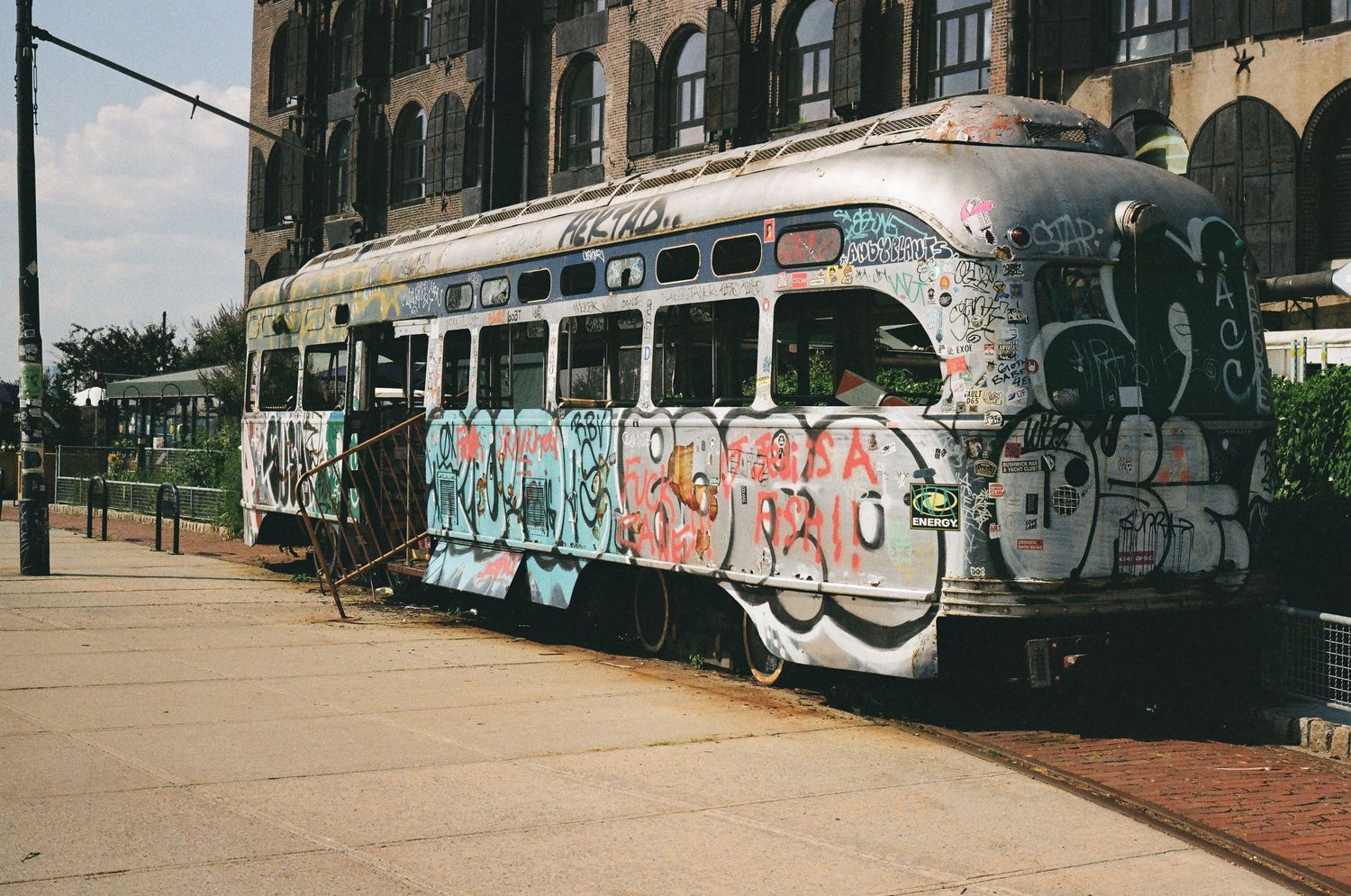 A vintage trolley car covered in colorful graffiti parked on a city street near a brick building.