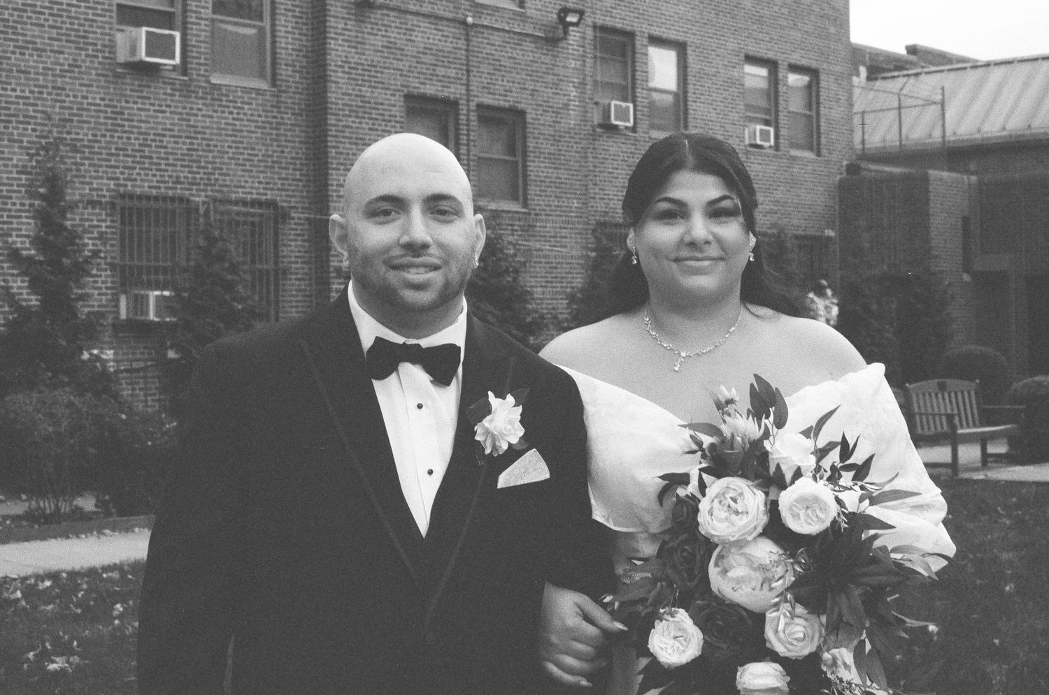 Black and white wedding portrait of a bald groom in a tuxedo and bride holding a bouquet outdoors.