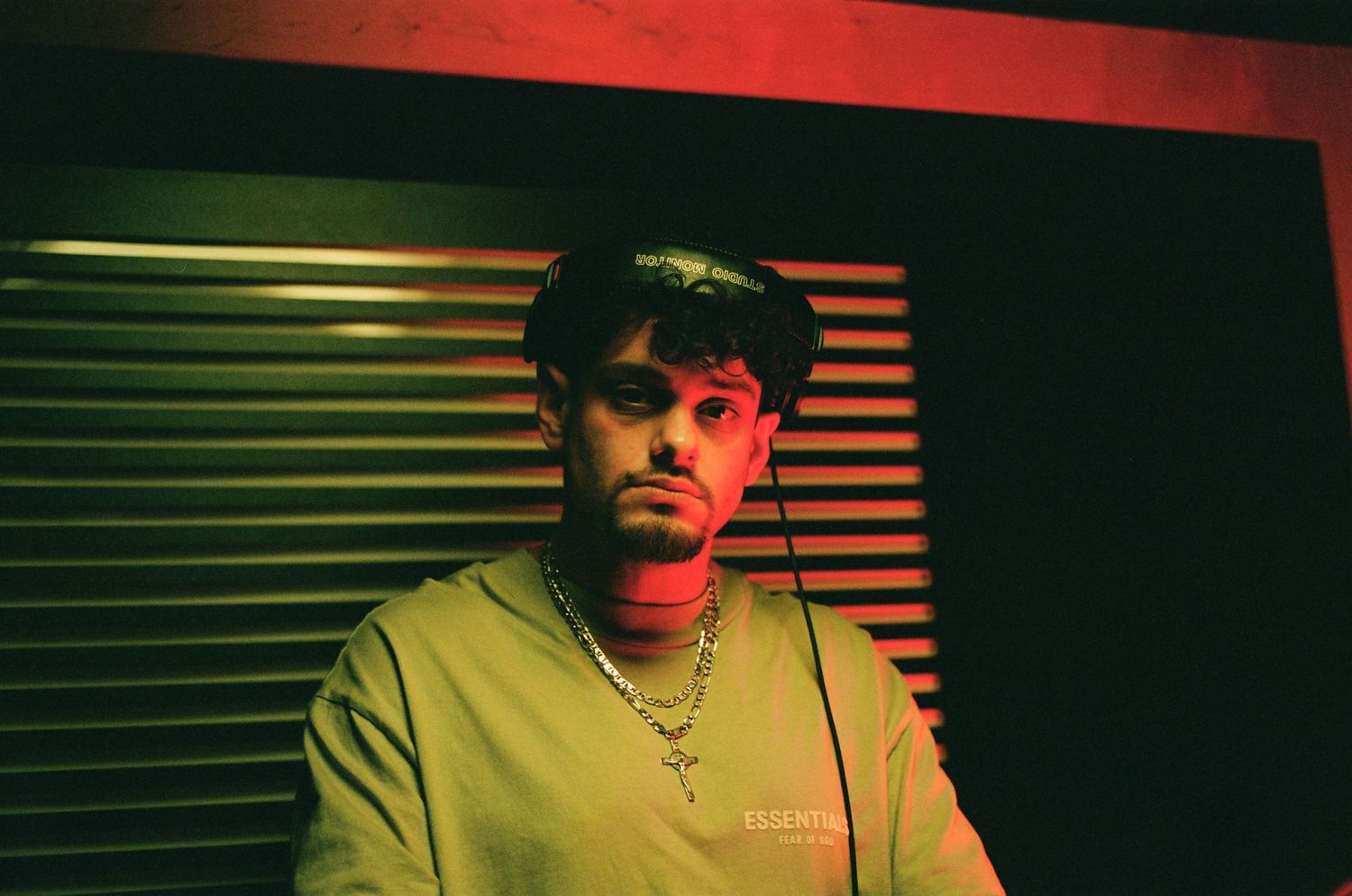 Young man with a chain necklace posing under dramatic red moody lighting in a dark room.