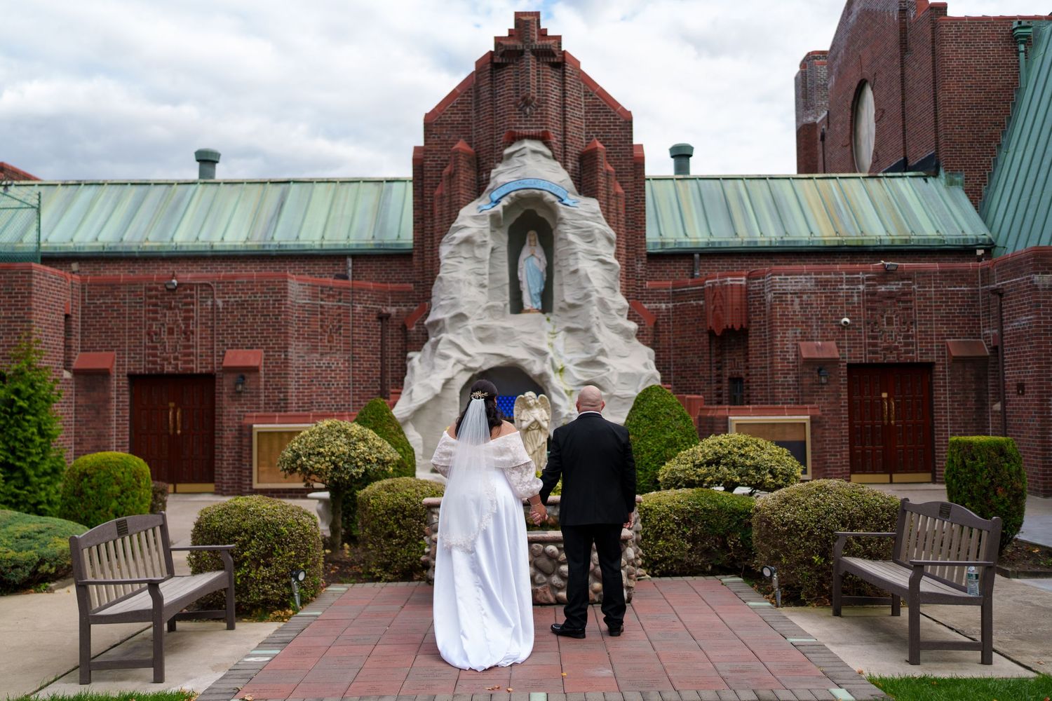 Bride and groom standing before a Marian grotto outside a red brick Catholic church.