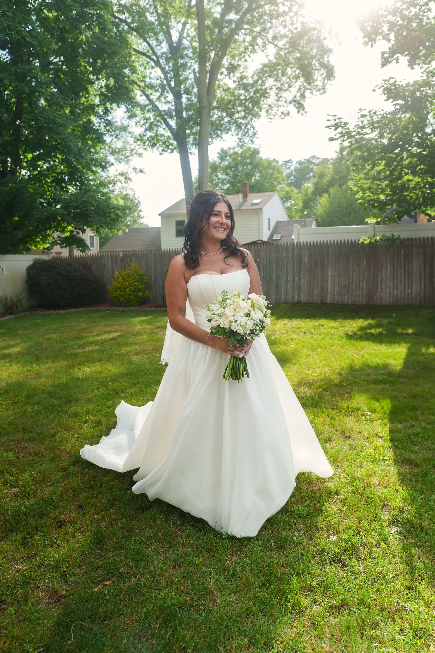 Bride in strapless white ball gown holding white bouquet standing in sunny backyard garden.