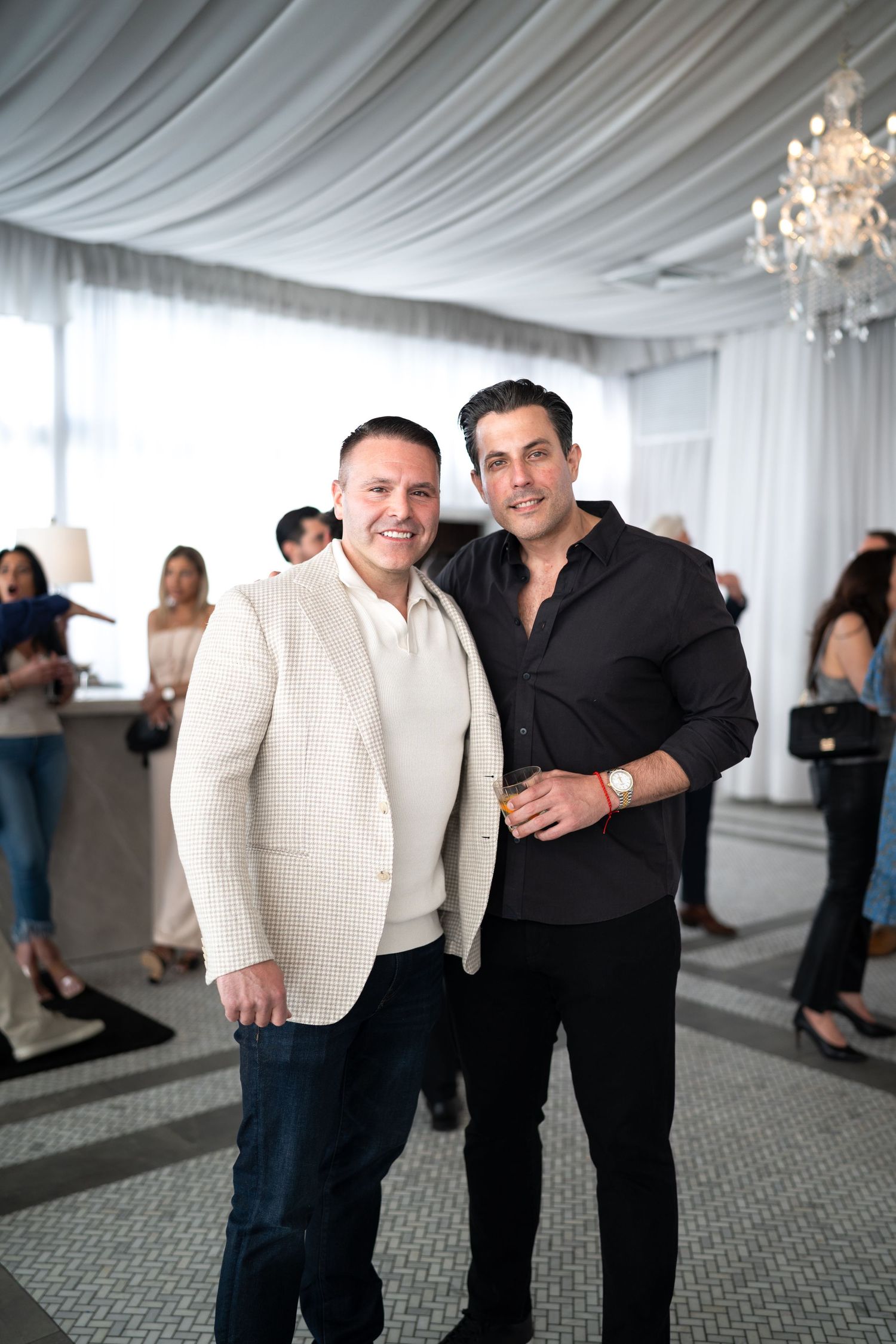 Two men smiling together at an upscale indoor event with chandelier and draped white ceiling.