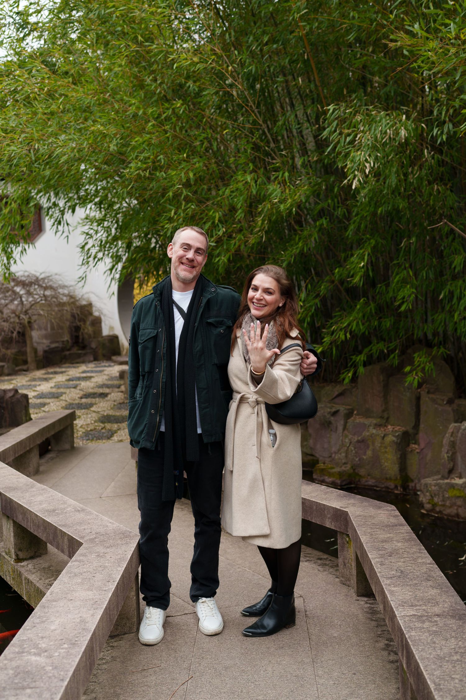Happy couple posing on a stone bridge showing off an engagement ring with bamboo trees behind them.