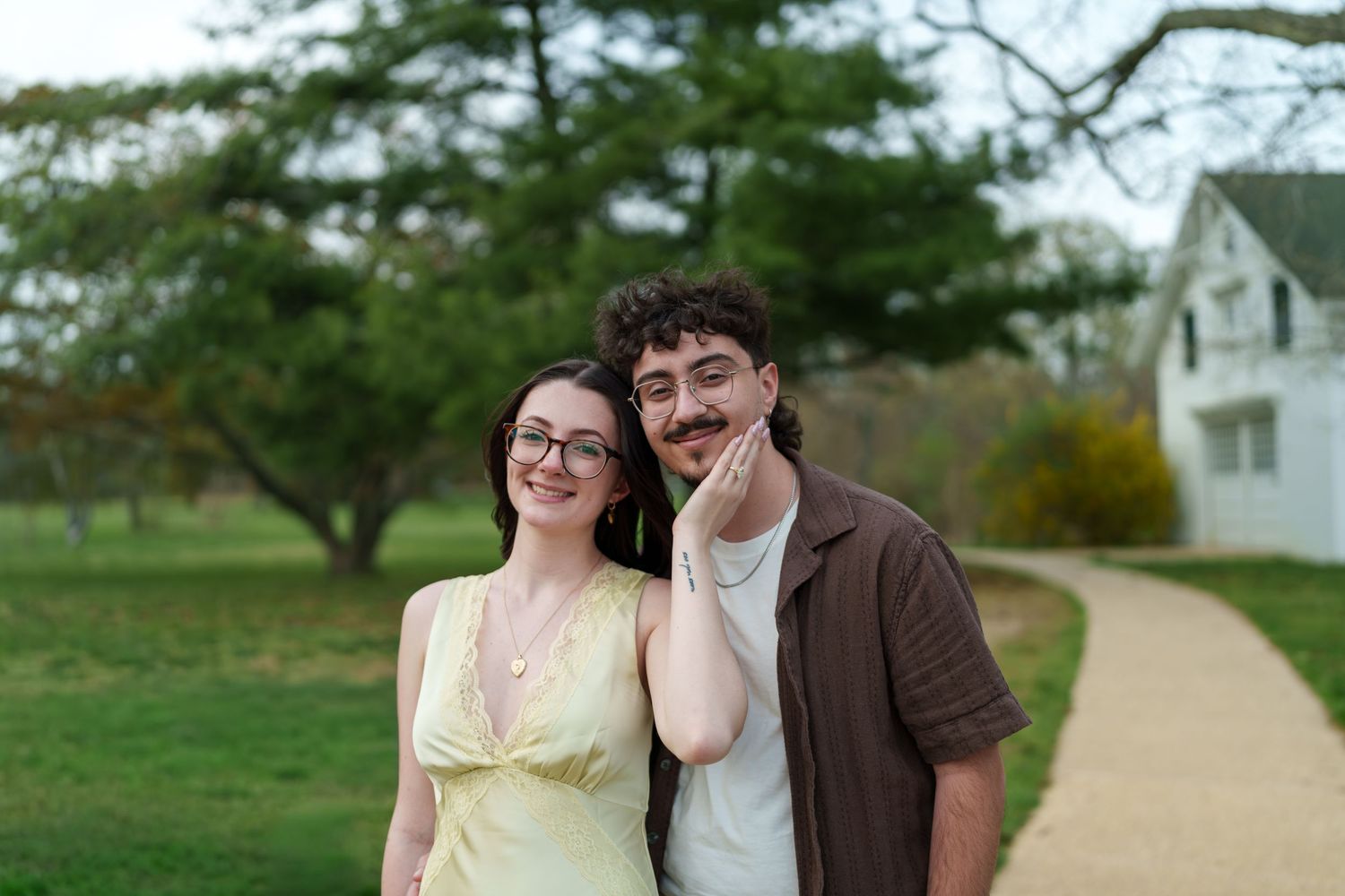 Smiling engaged couple posing together on a park path, woman in yellow dress and man in brown shirt.