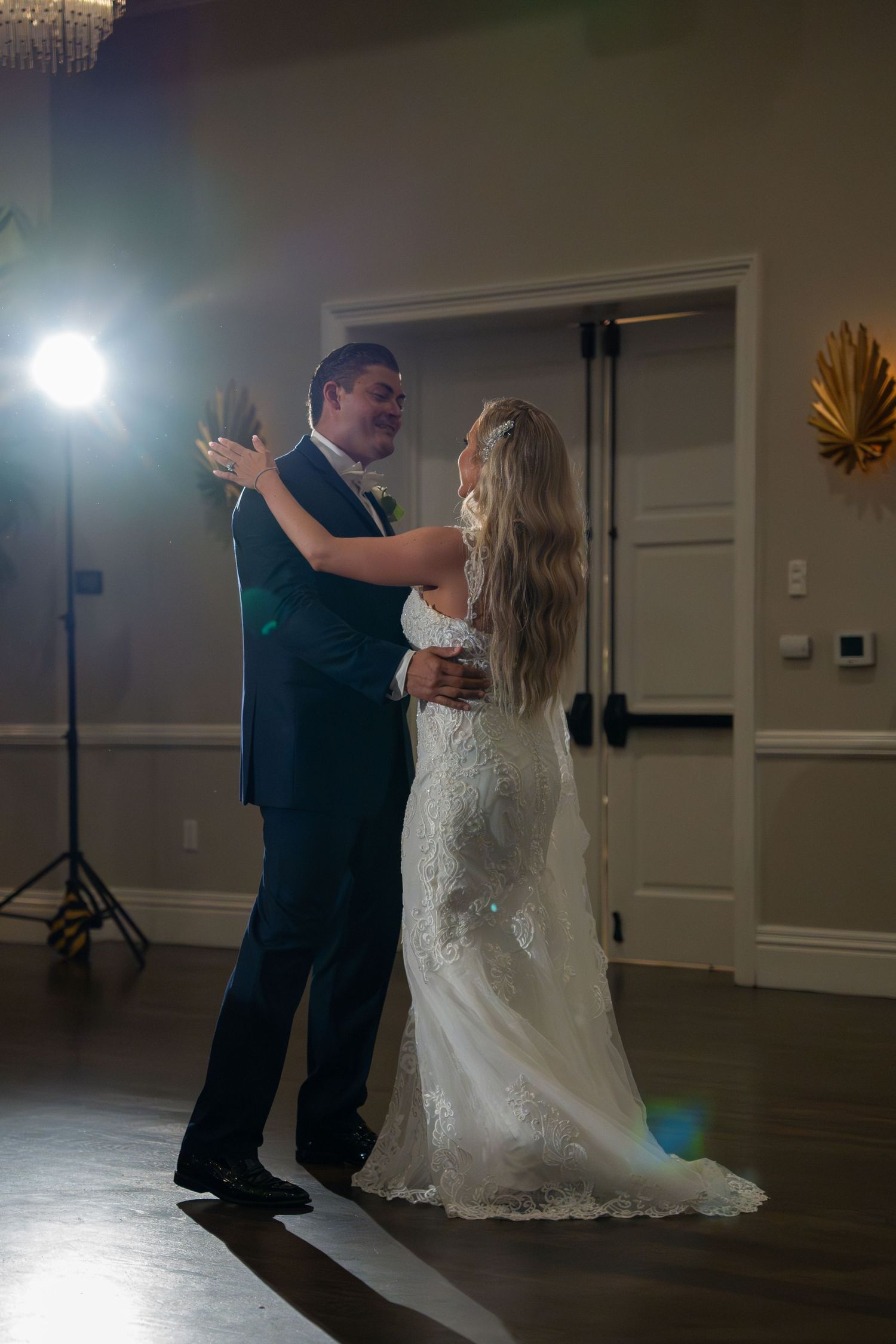 Bride in lace gown and groom in navy suit share first dance in softly lit ballroom.