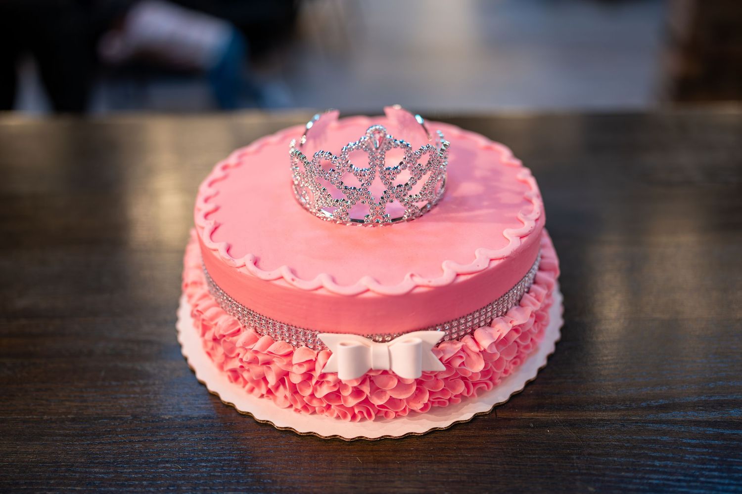 Pink princess cake with silver tiara decoration and ruffle frosting on wooden table.