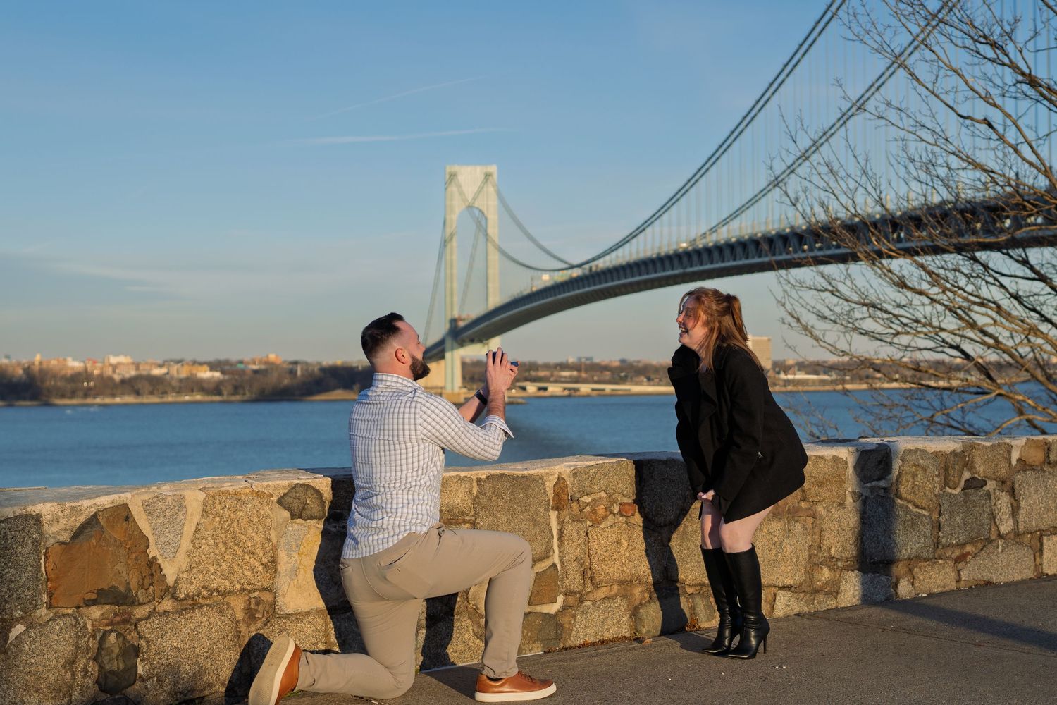 Romantic marriage proposal moment with man kneeling before woman at scenic Verrazano Bridge waterfront in New York.