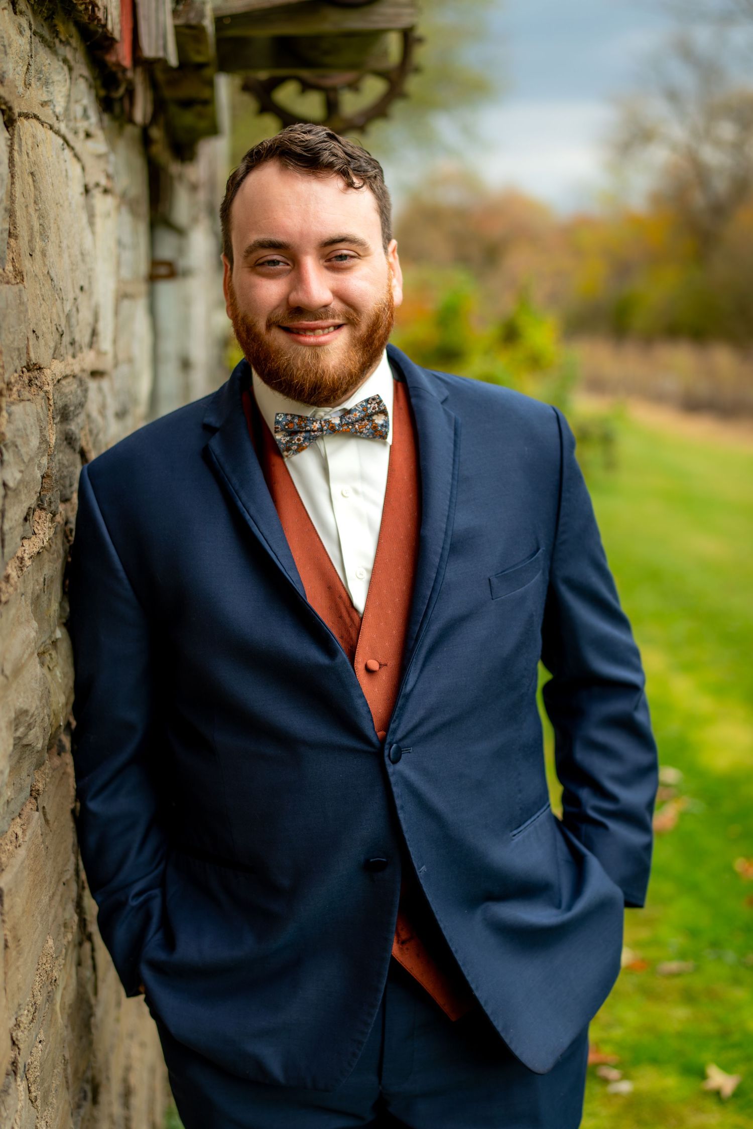 Handsome groom in navy suit with rust vest and bow tie poses against stone wall outdoors.