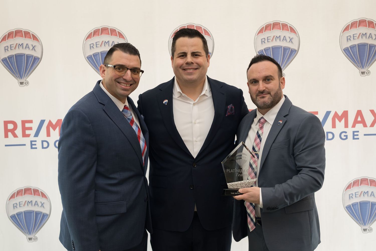 Three RE/MAX Edge real estate professionals smile together holding a glass trophy at awards event.