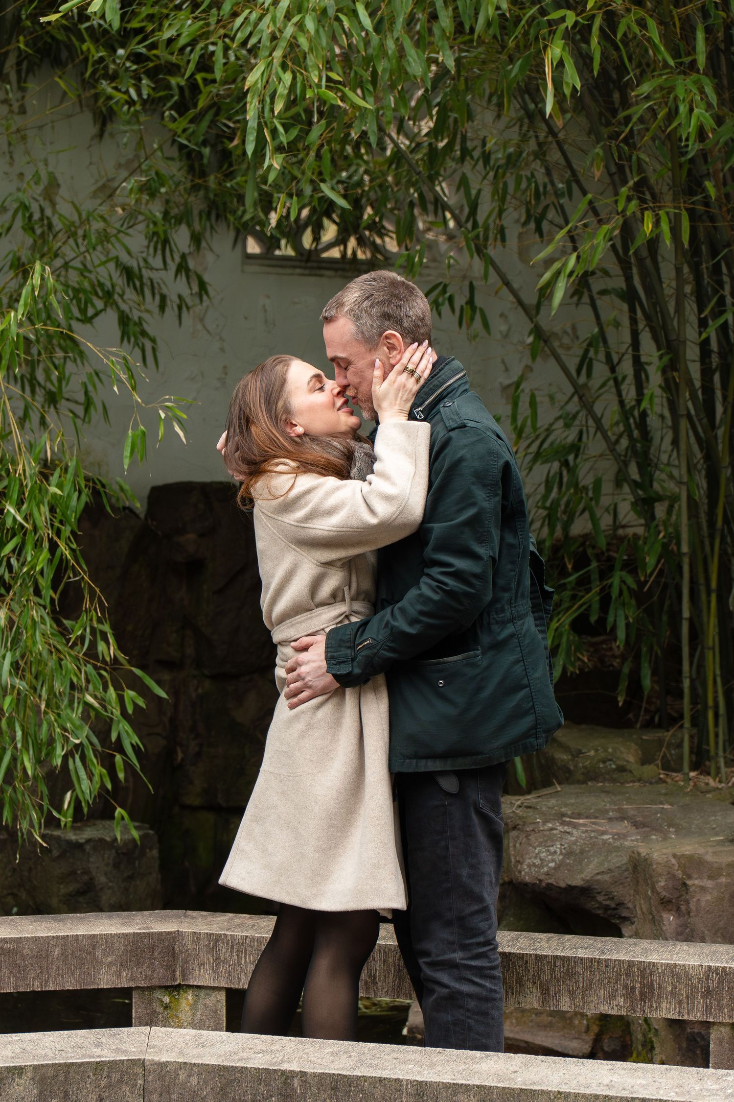 Couple sharing a tender kiss on a stone bridge surrounded by lush green bamboo in a garden.