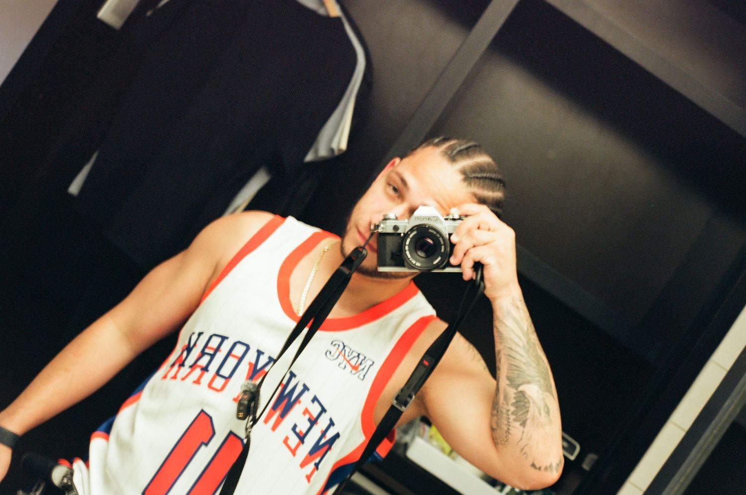 Person in a basketball jersey taking a mirror selfie with a film camera, showing tattoos.