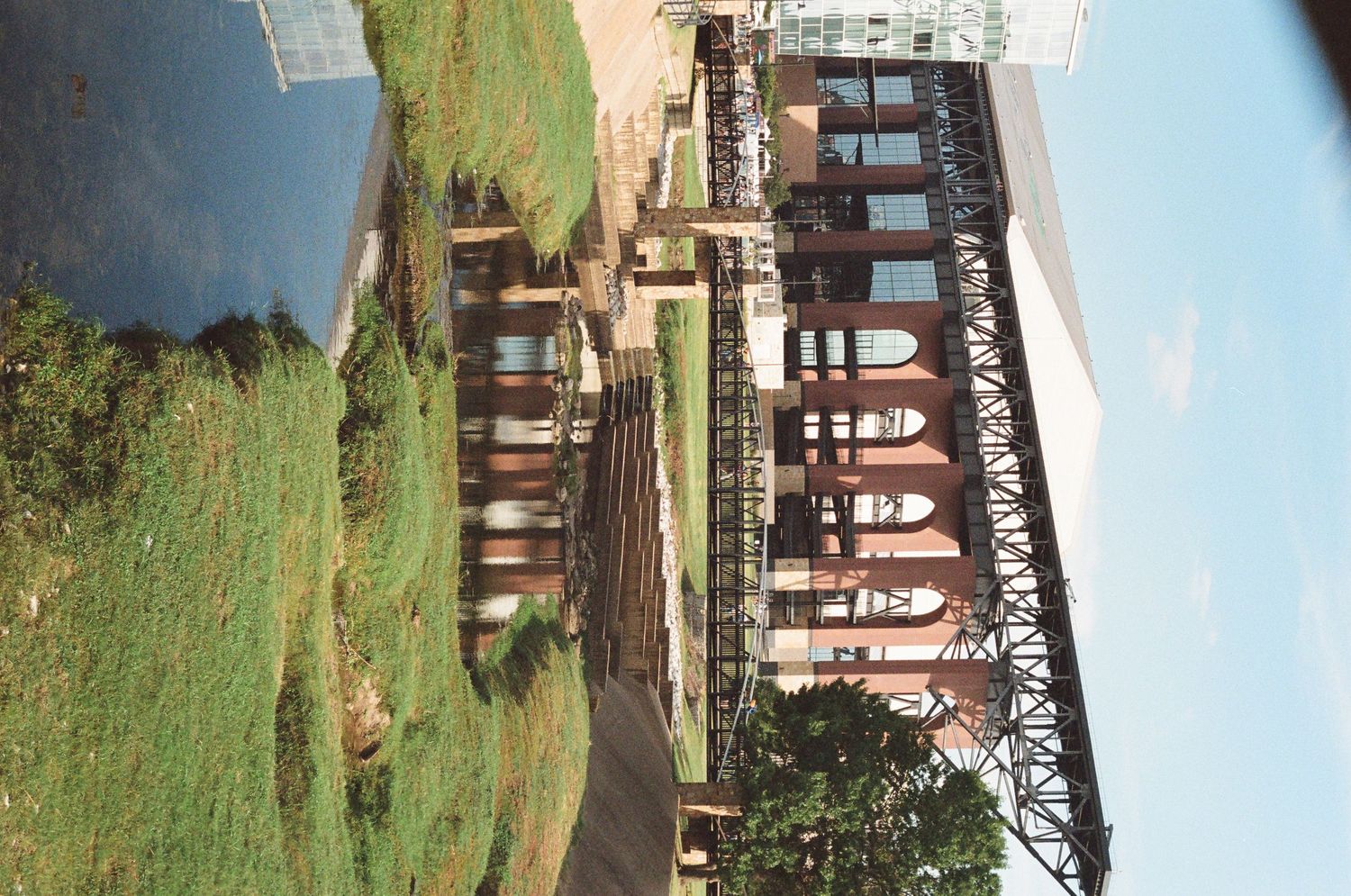 Historic red brick industrial building with a metal railway bridge structure beside a green lawn.
