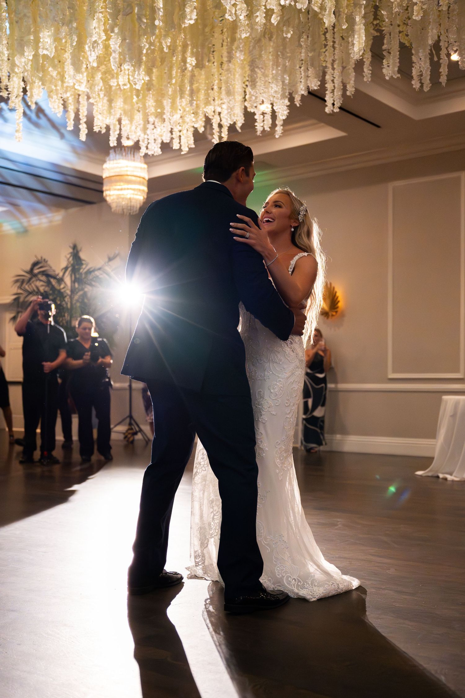 Bride in white lace gown laughs joyfully with groom during first dance at wedding reception.