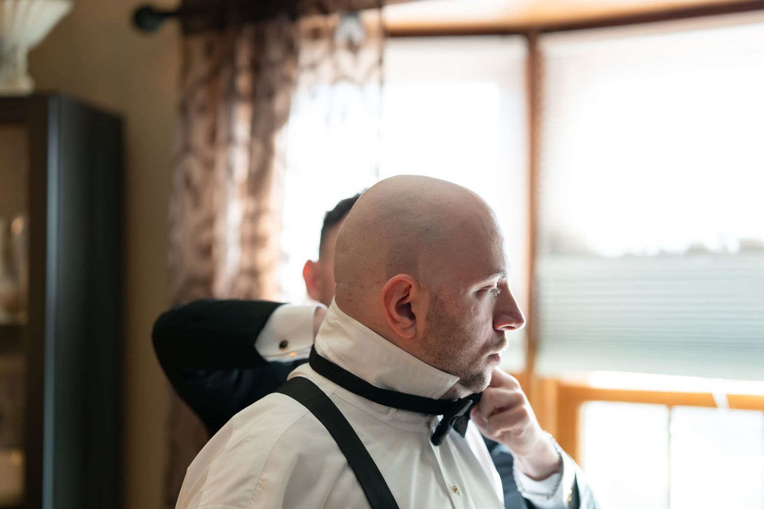 Bald groom adjusting his bow tie while getting ready, wearing white dress shirt and black suspenders indoors.