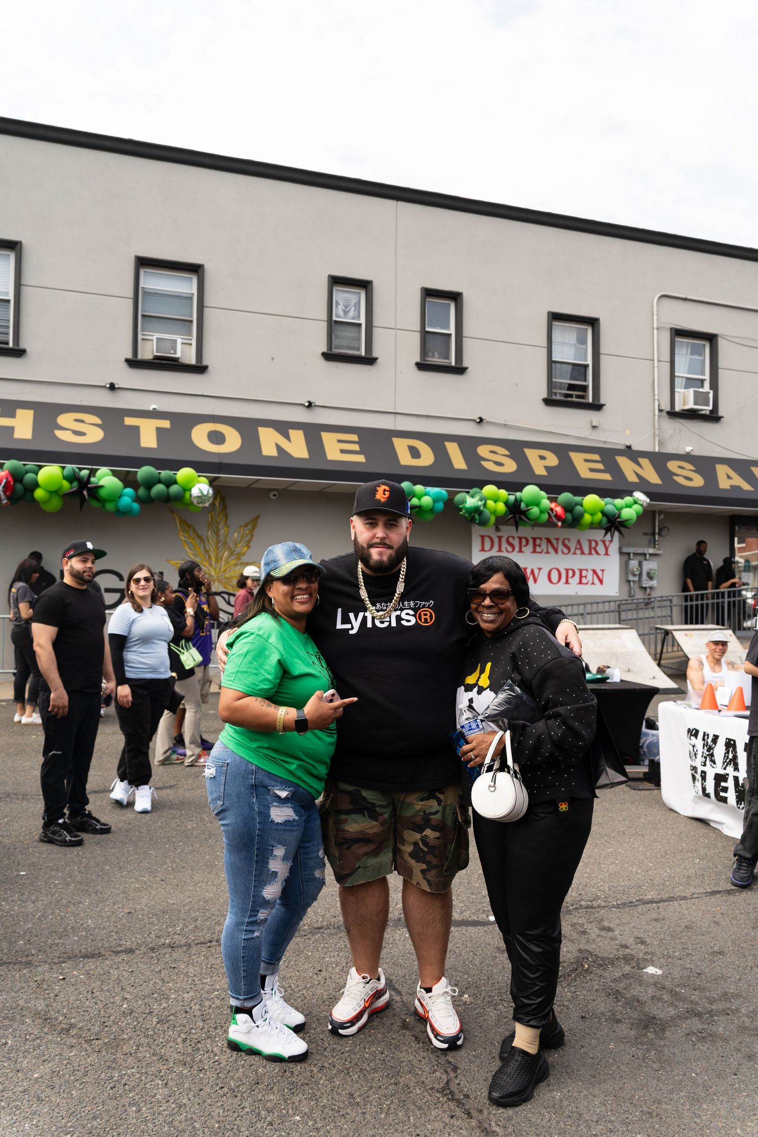 Three people posing together outside a cannabis dispensary with green balloon decorations at grand opening.