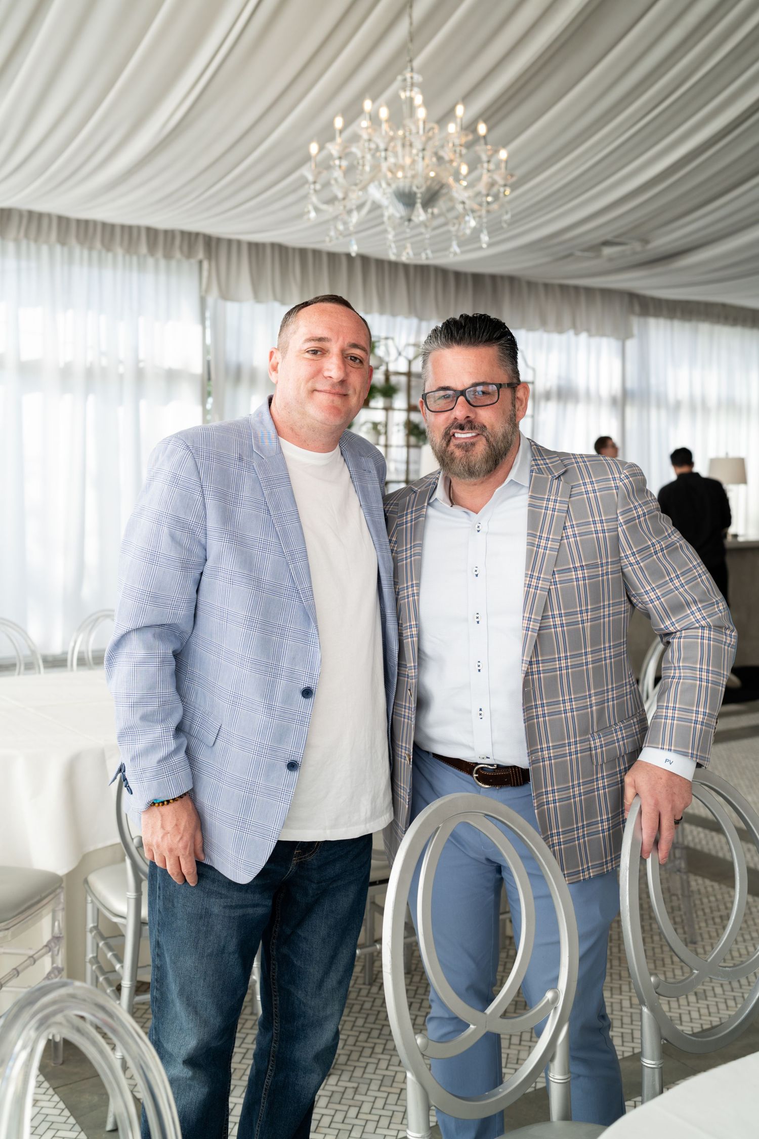 Two men in blazers pose together in an elegant white event venue with crystal chandelier.
