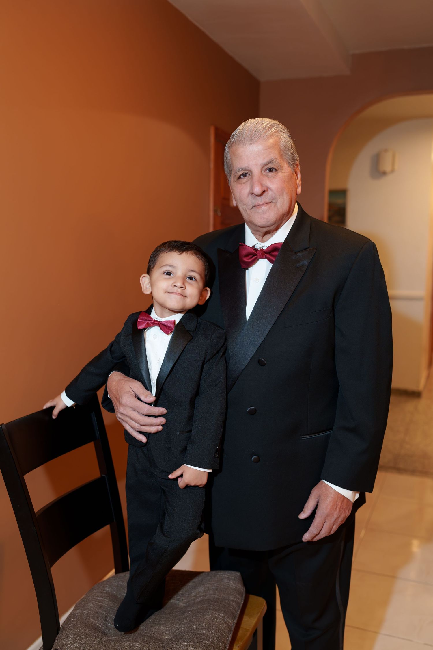 Grandfather and grandson both dressed in matching black tuxedos with burgundy bow ties posing together.