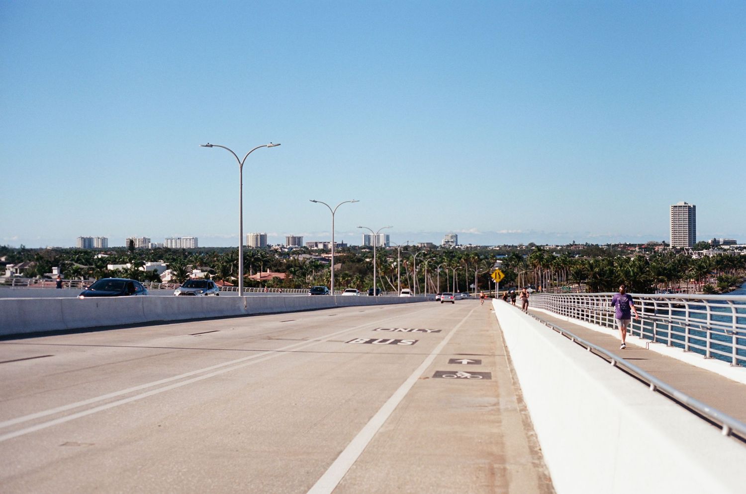 Wide bridge road with bike lane and pedestrians, overlooking a coastal city skyline on a clear day.