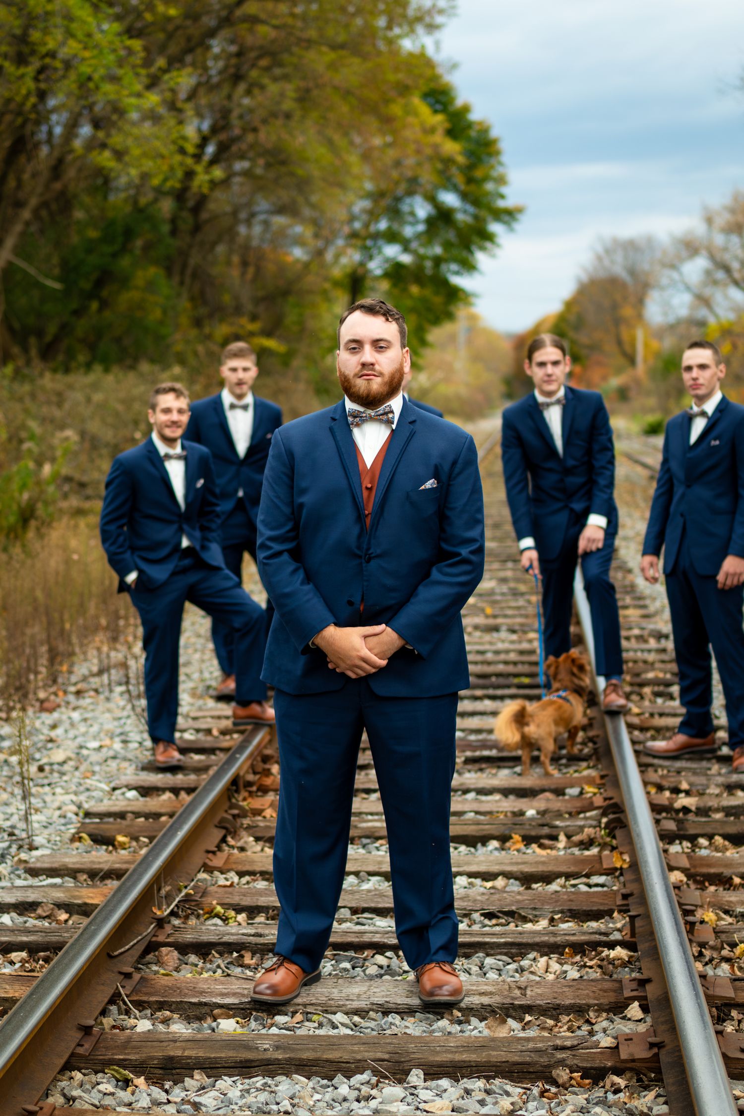 Groom and groomsmen in navy suits pose on railroad tracks surrounded by autumn foliage with dog.