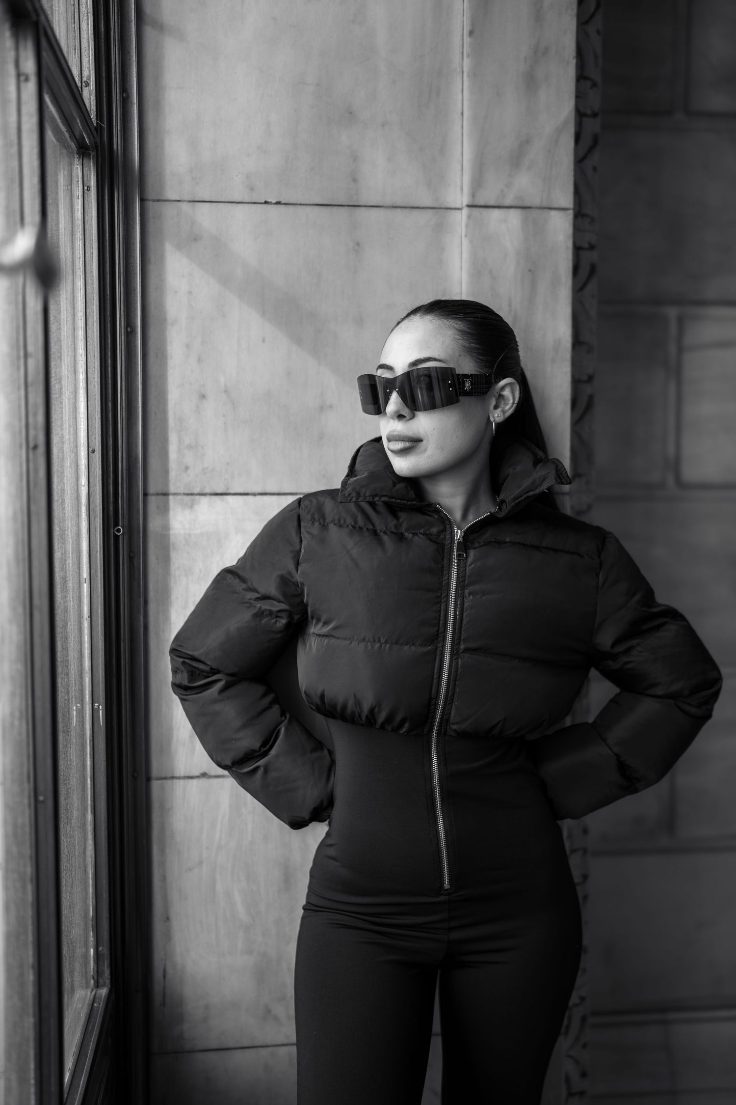 Black and white photo of woman in puffer jacket and sunglasses posing by window and stone wall.