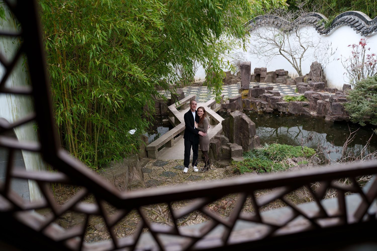 Couple posing by a pond and rock garden framed by an intricate decorative lattice window in a Chinese garden.
