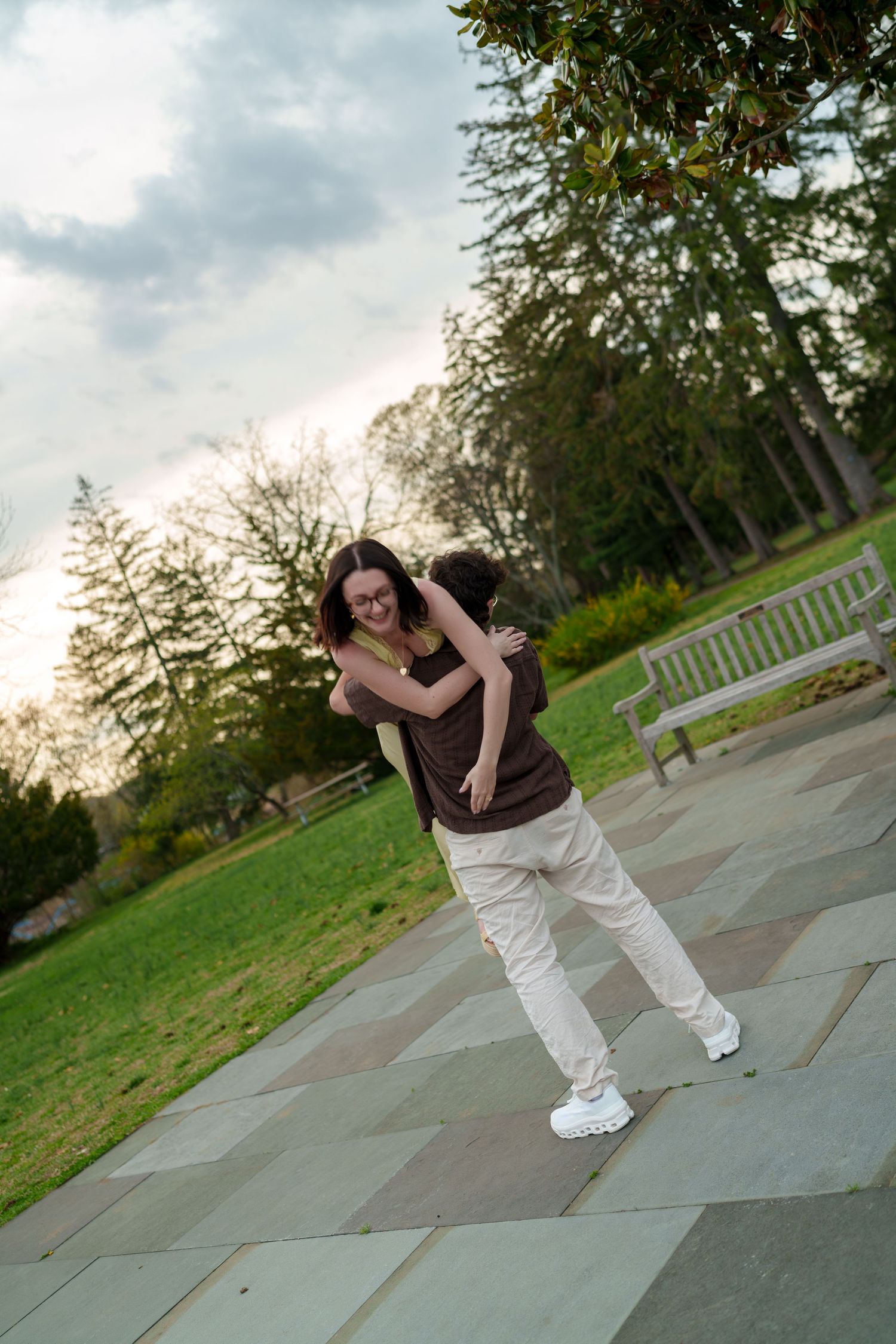 Man lifting woman in a joyful hug on a park pathway with green lawn and bench behind them.