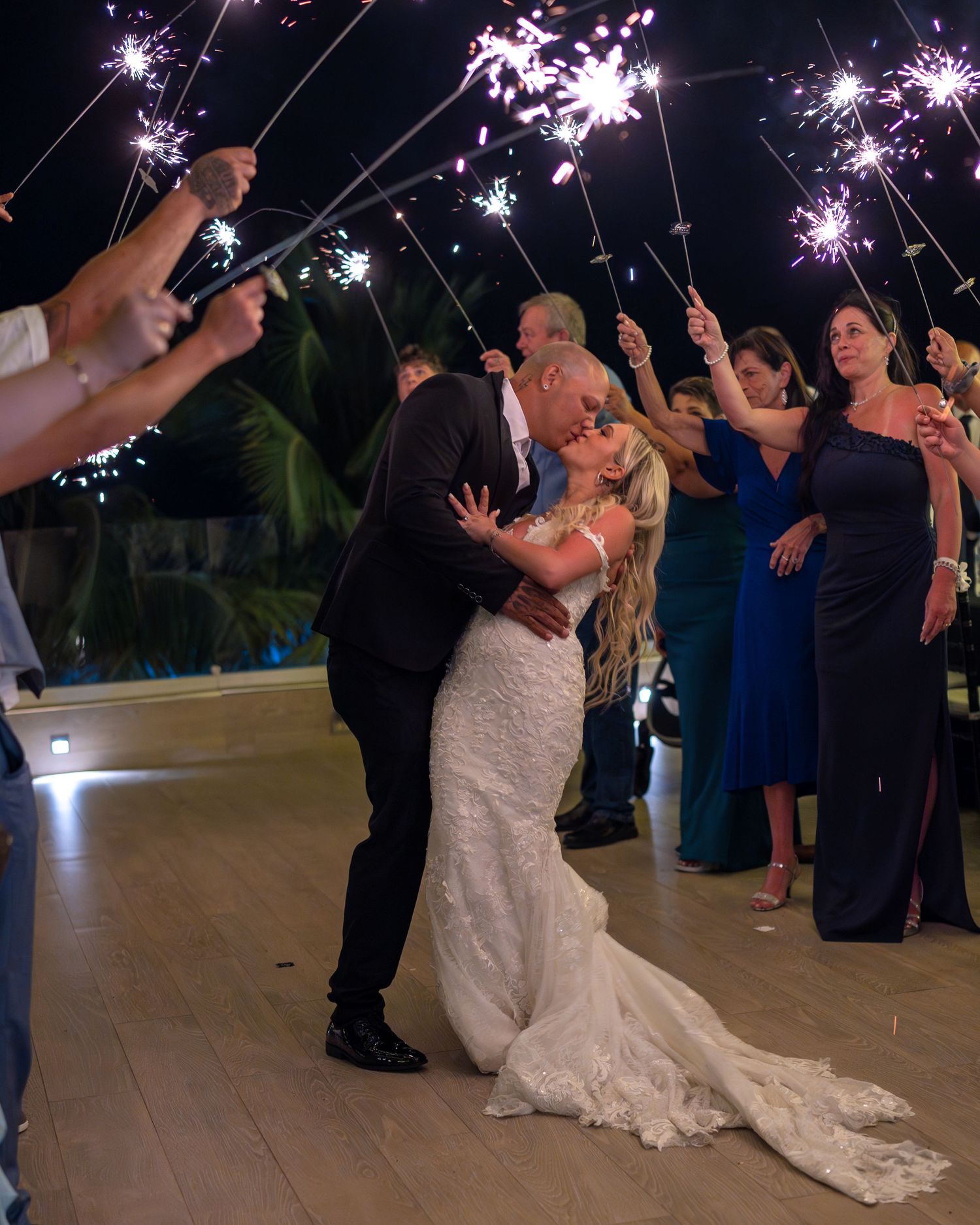 Groom dips bride in passionate kiss surrounded by guests holding sparklers on outdoor wedding dance floor.