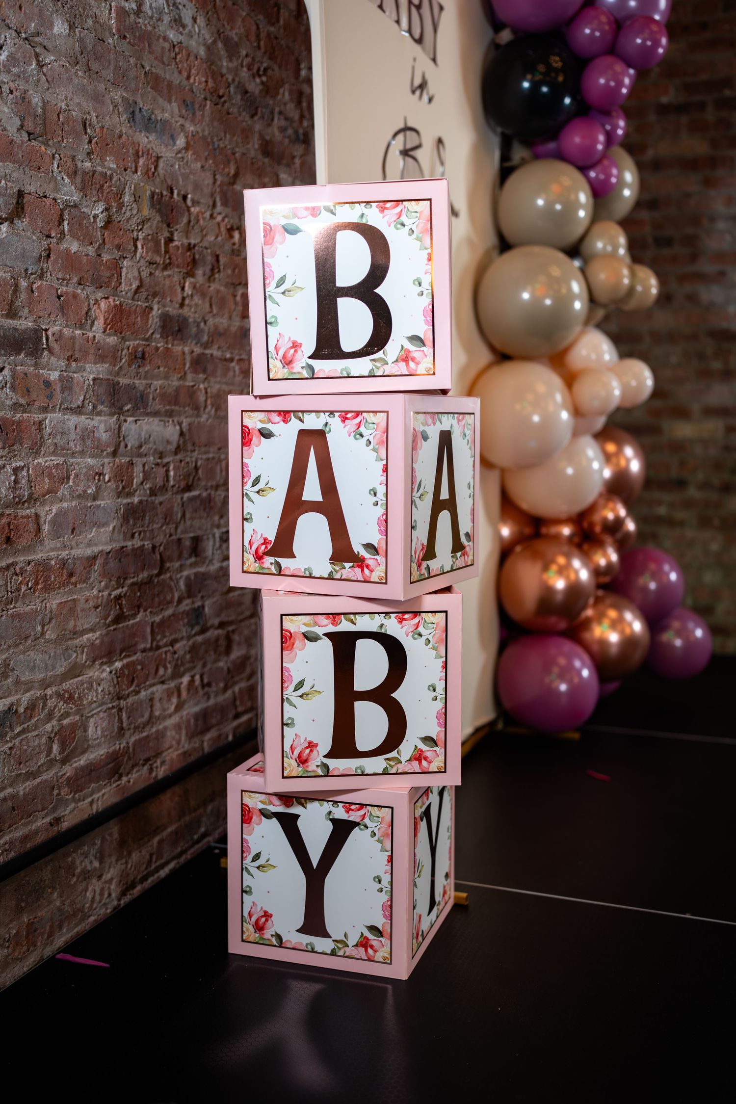 Pink floral BABY letter blocks stacked vertically beside colorful balloon garland at shower.