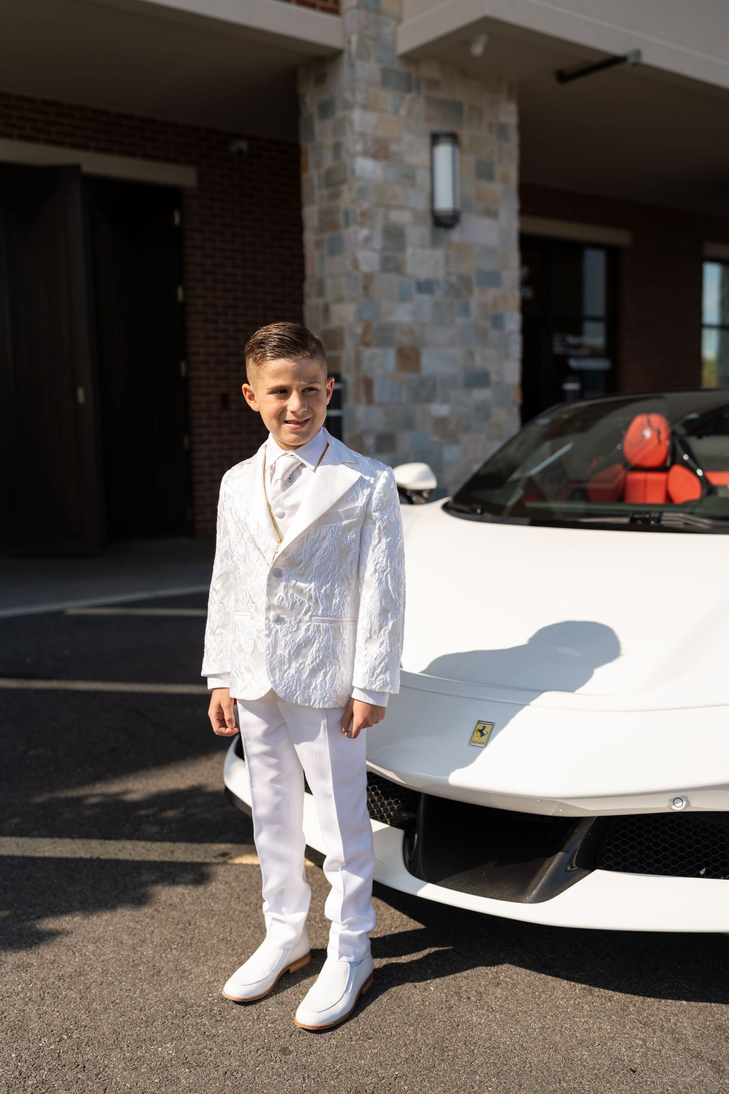 Young boy in white suit posing proudly in front of a white Ferrari sports car.