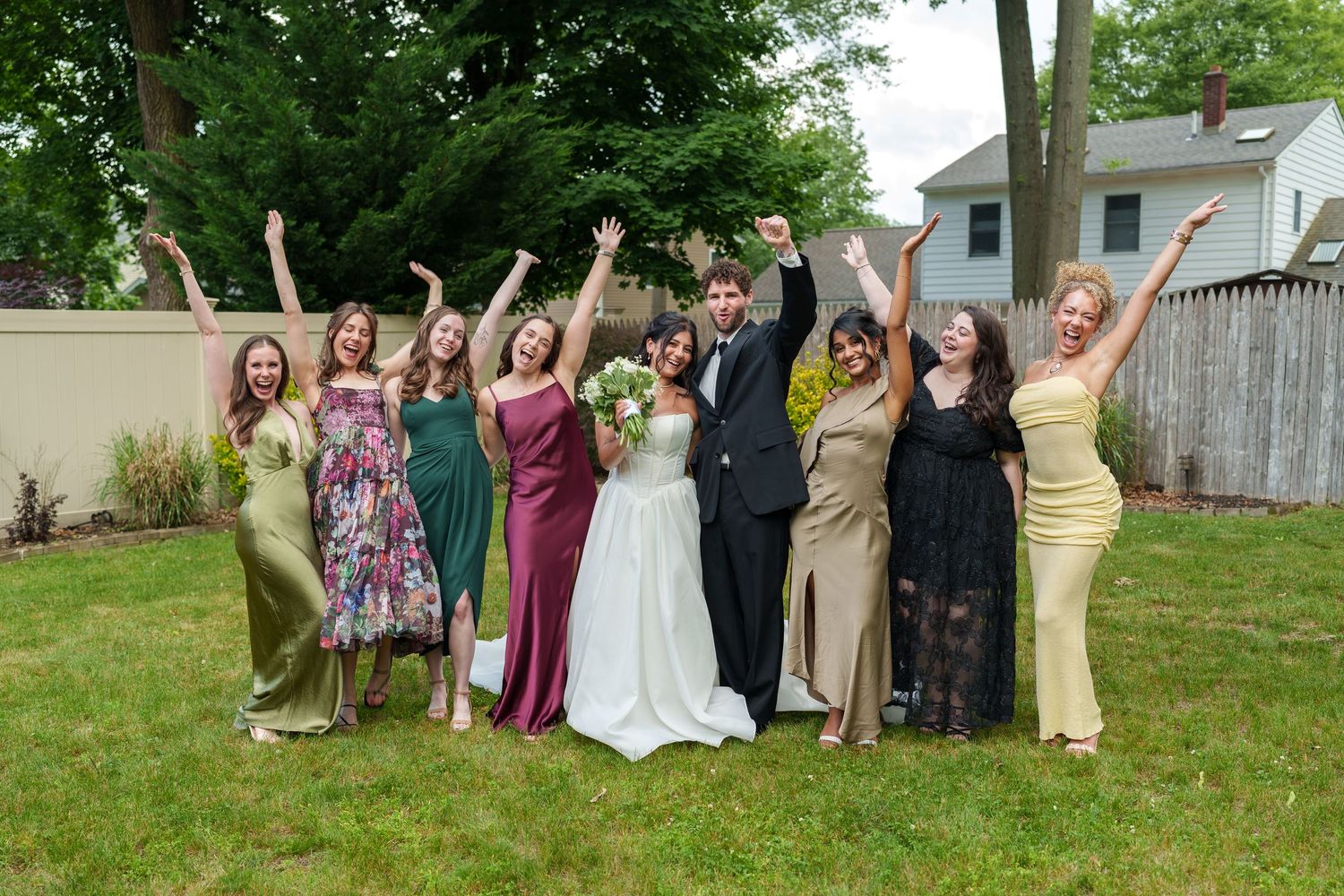 Joyful wedding party group photo on a lawn, bride and groom surrounded by bridesmaids in colorful dresses cheering.