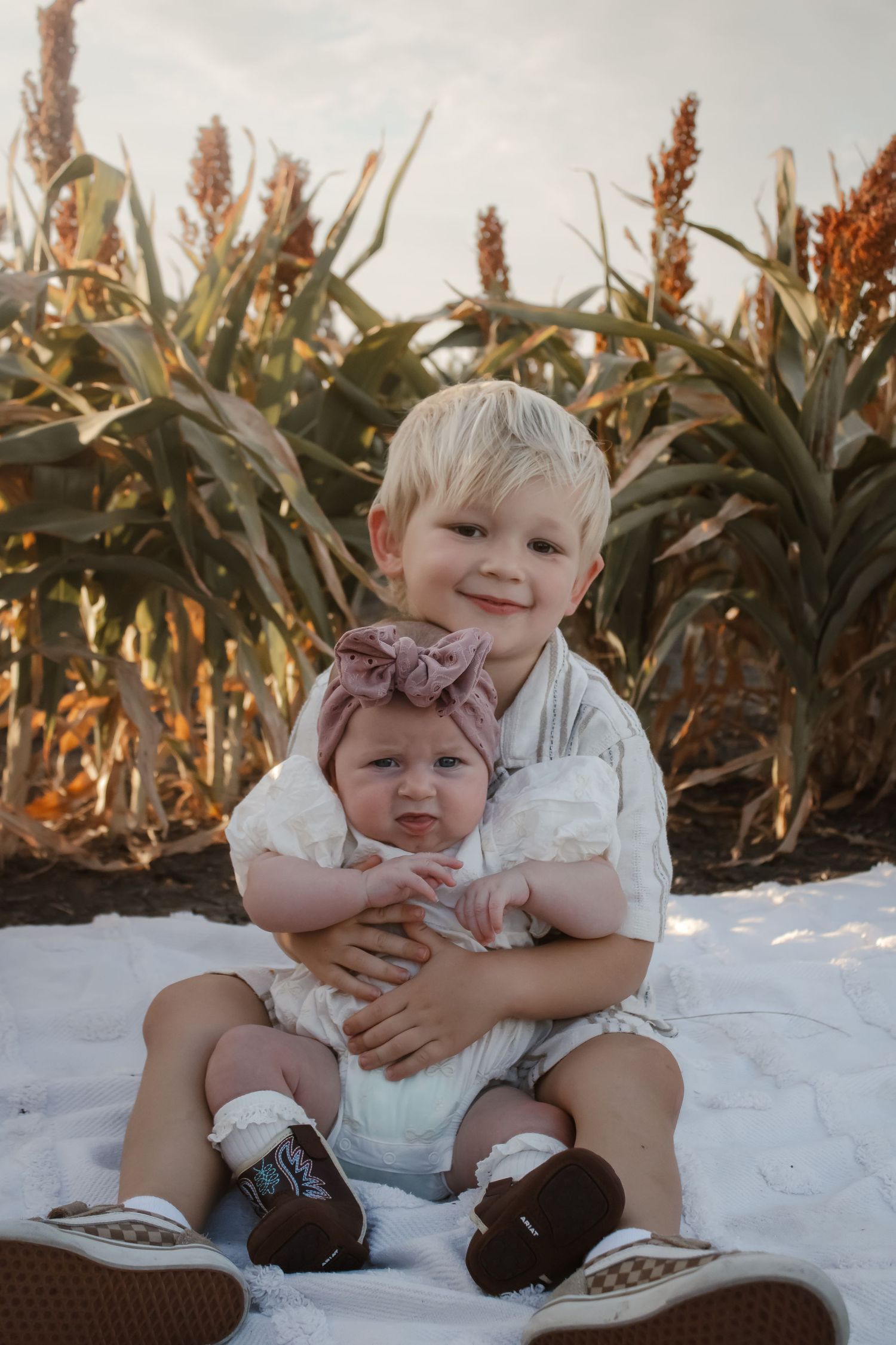 Two children sitting together on a white blanket in front of a sorghum field at sunset.