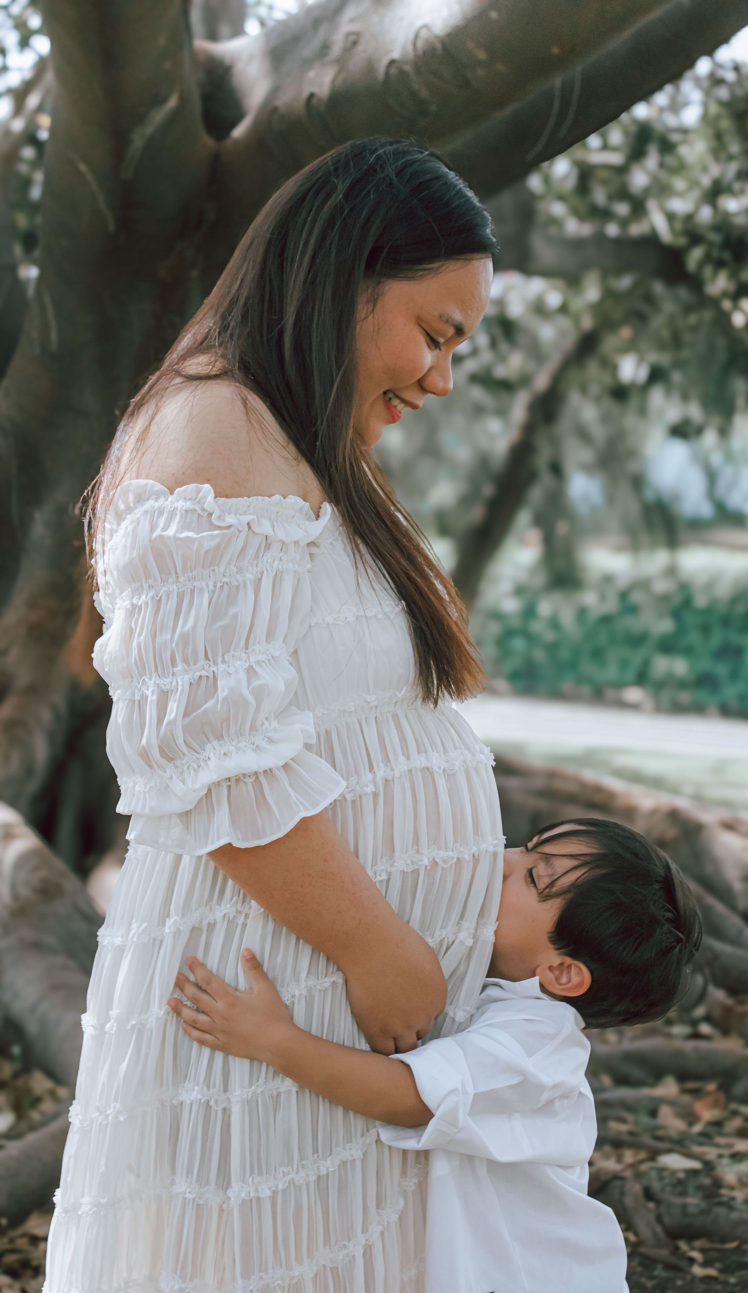 Mother and child in white dresses share tender moments under blooming trees during outdoor photo session.
