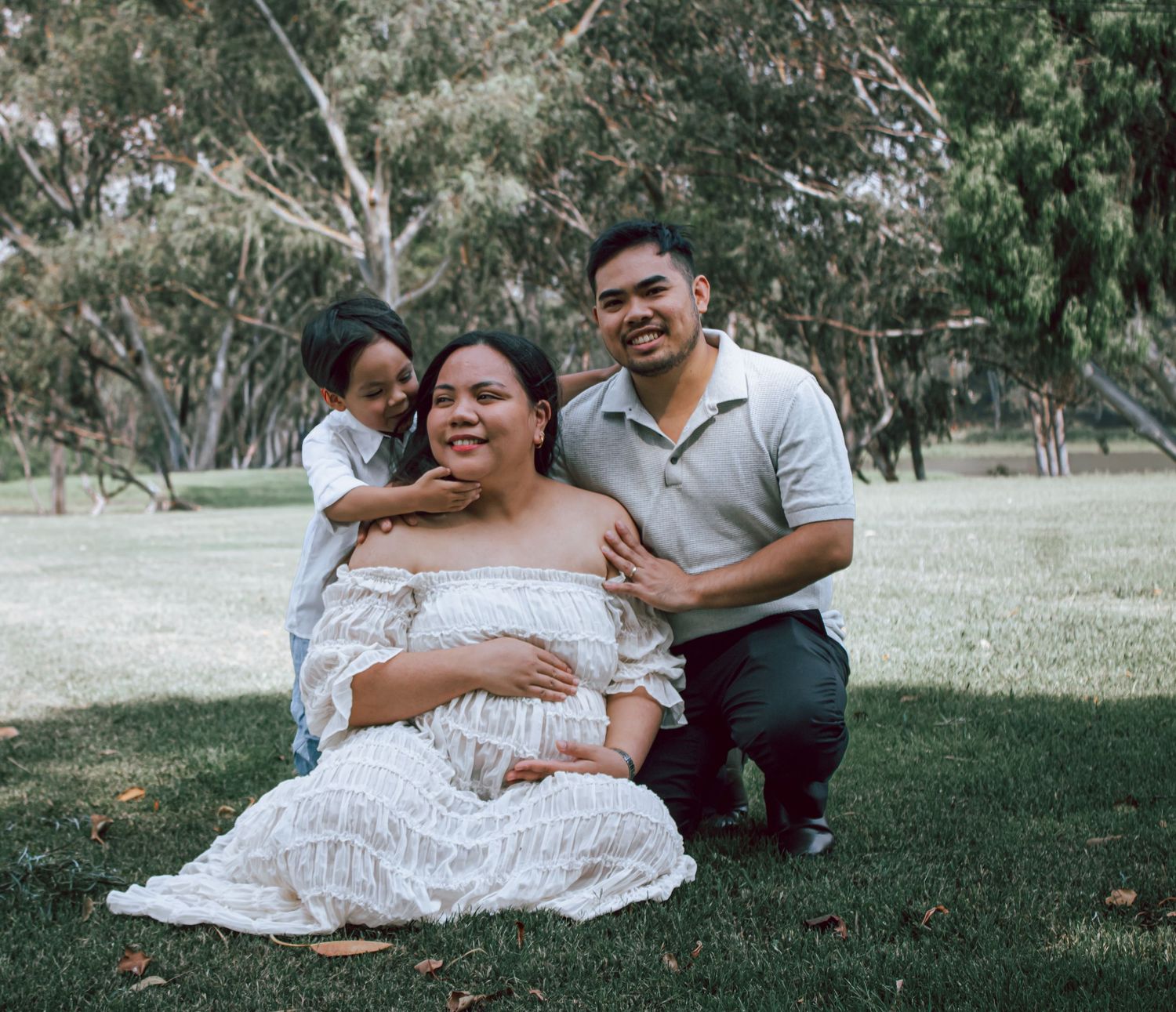 A couple sits together on grass during a romantic outdoor maternity photoshoot in a peaceful park setting.