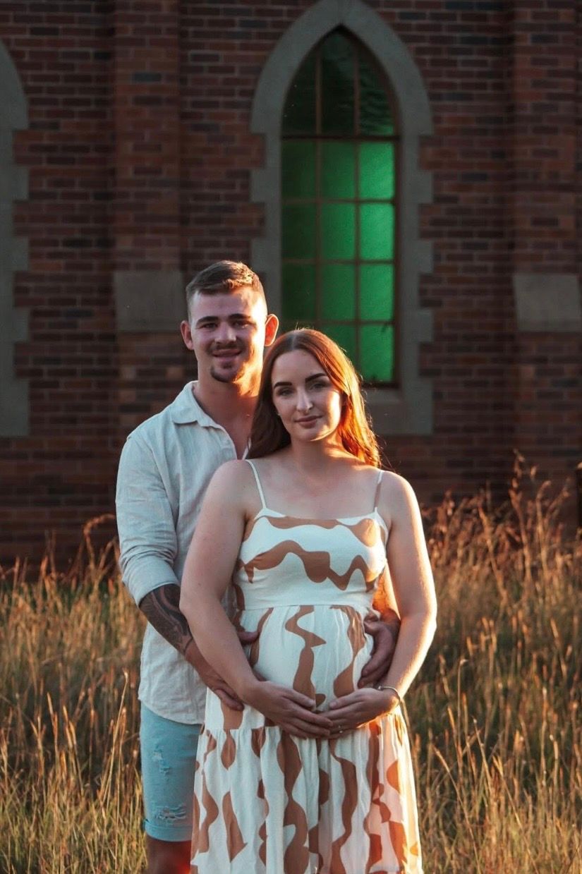 A couple embraces in a golden field at sunset with a brick building and green-lit windows in the background.