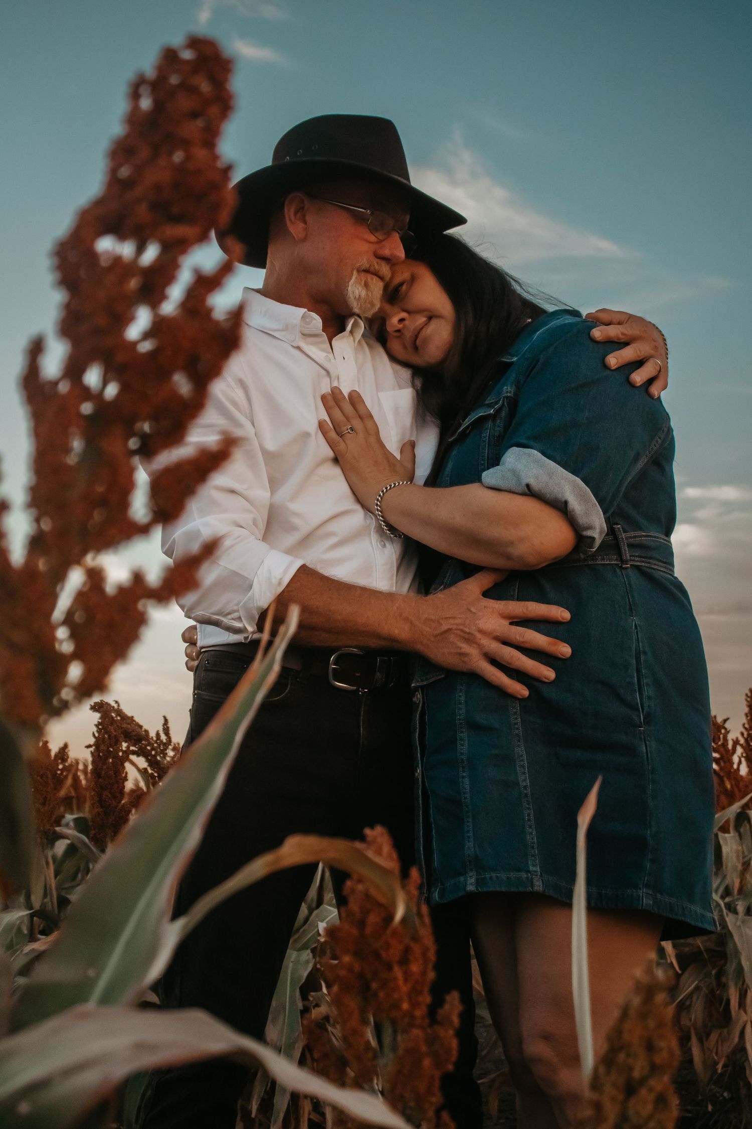 A romantic couple embraces in a field at sunset, with tall red sorghum plants in the background.